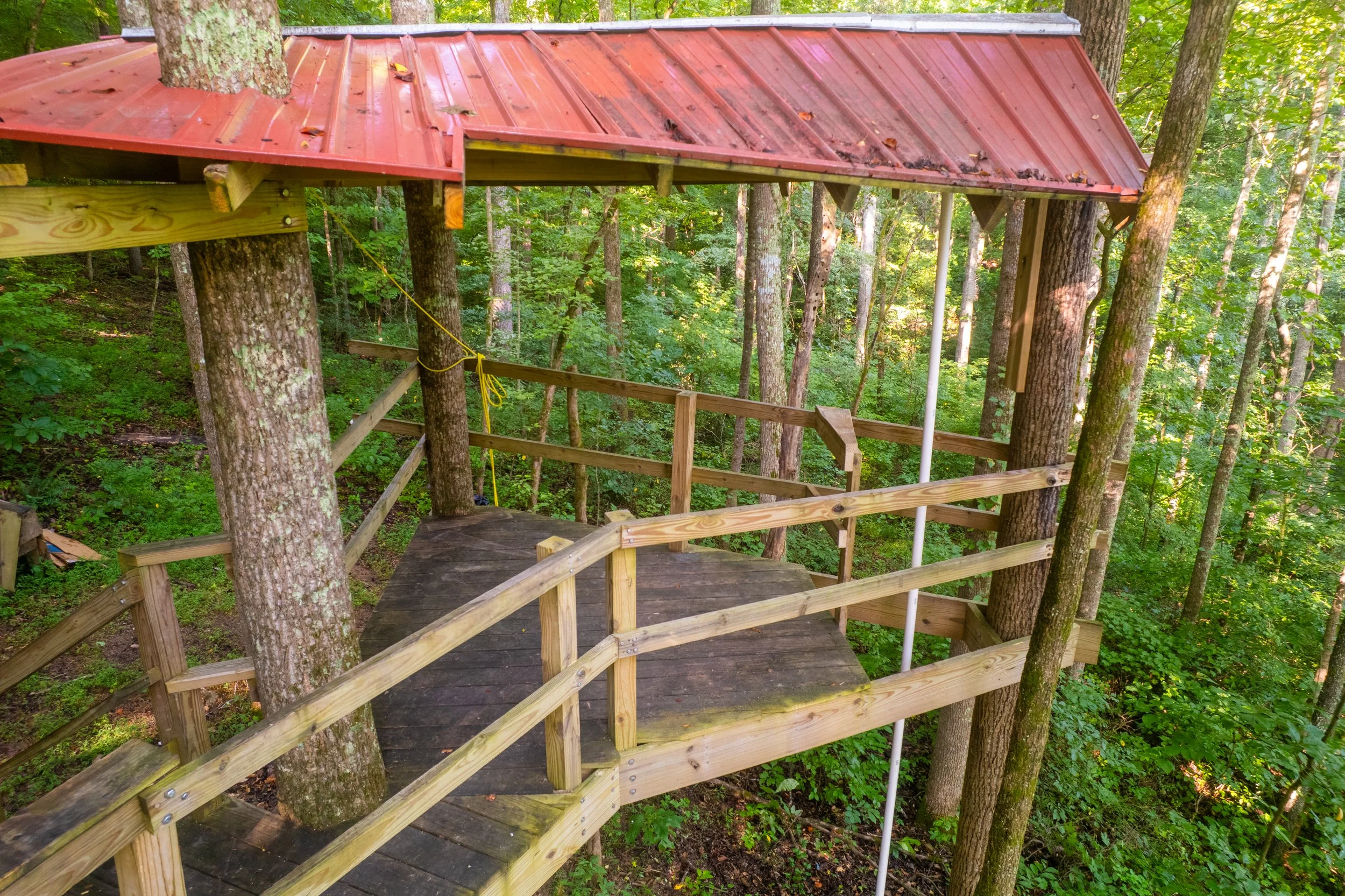 Treehouse with a red metal roof, wooden platform, and safety railings, built among trees in a forest.