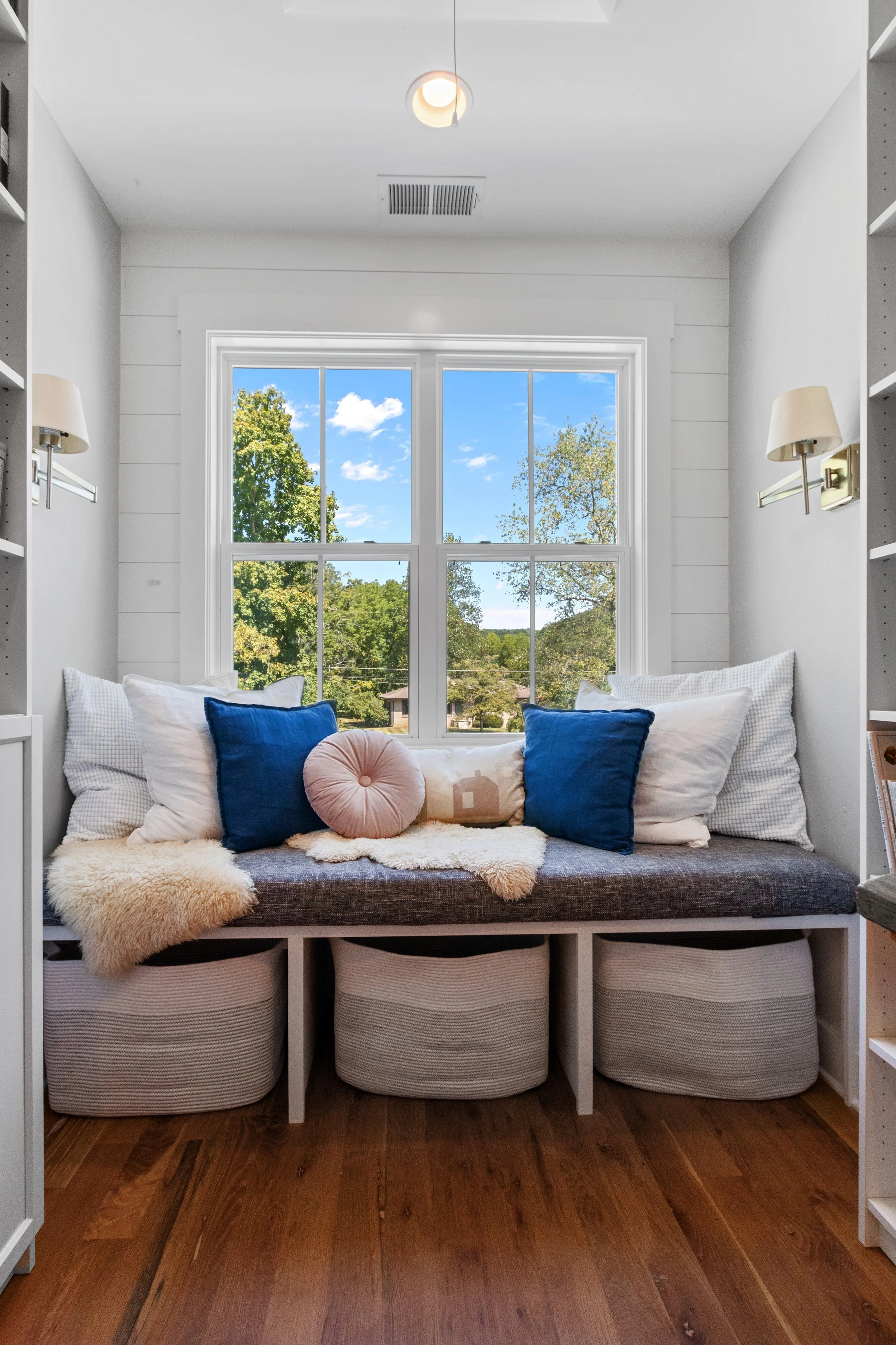 A cozy window nook with a cushioned bench, decorative pillows, and a view of trees and a blue sky through a large window.