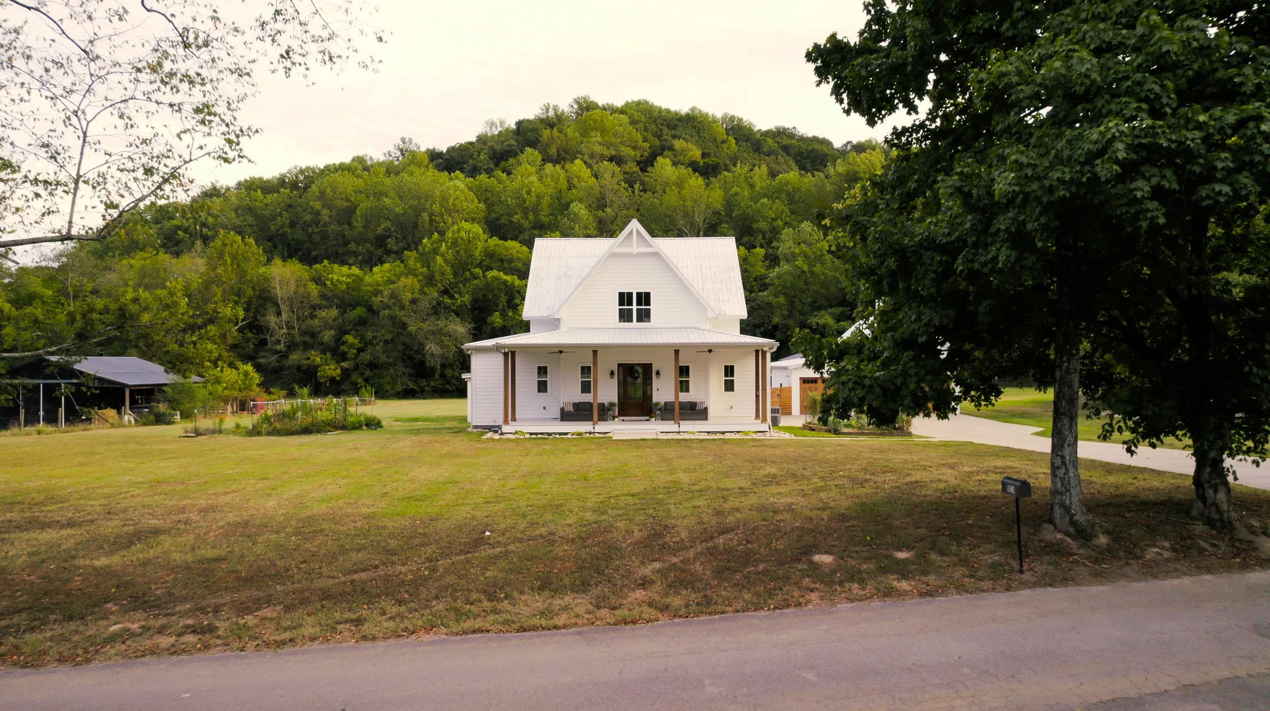A white house with a covered front porch and dark furniture, situated in a green landscape with trees and a hill in the background.