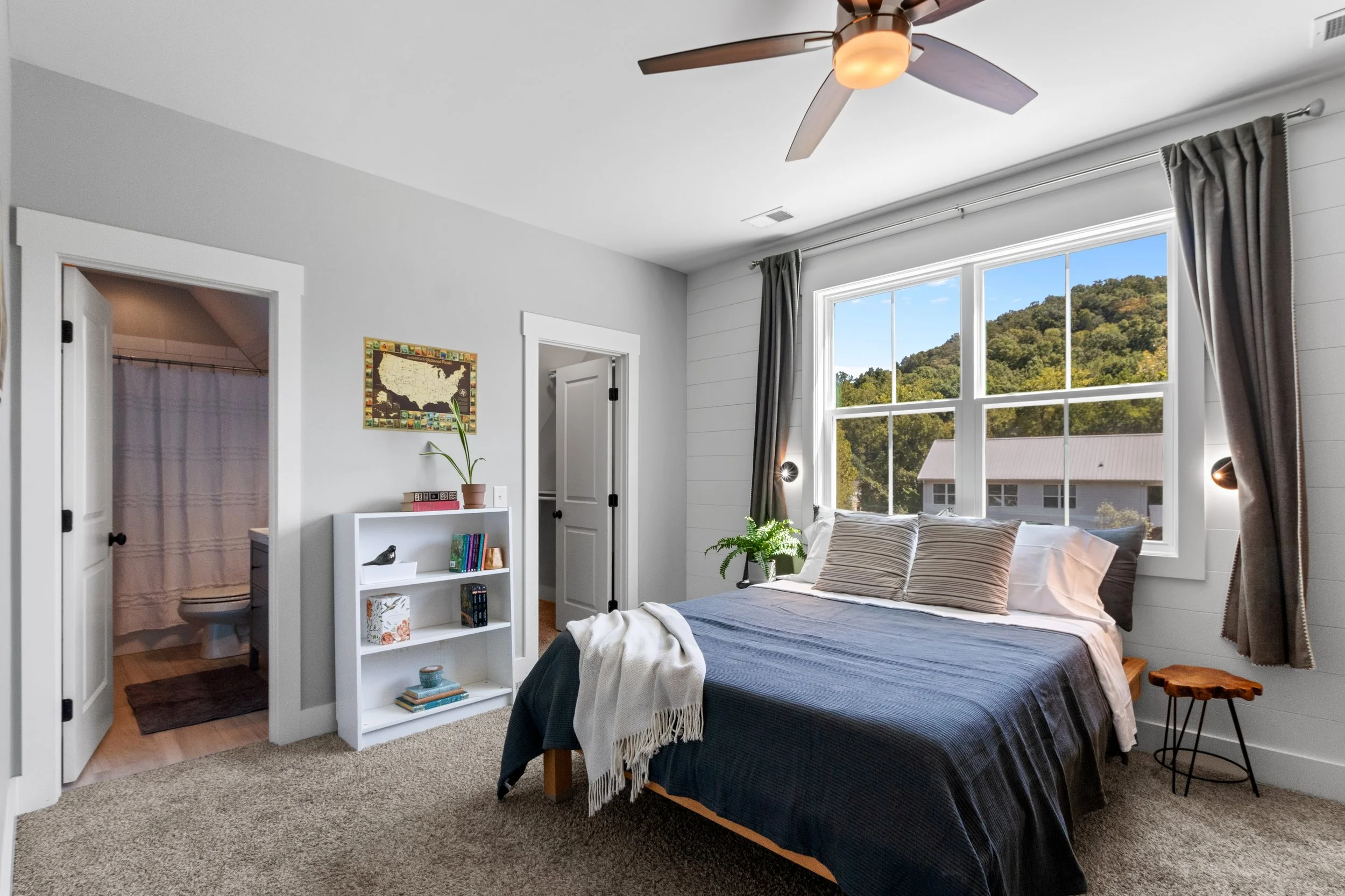 Bright bedroom with large window showing a green hill, bed with striped pillows and dark blue cover, ceiling fan, bookshelf, and an adjoining bathroom.