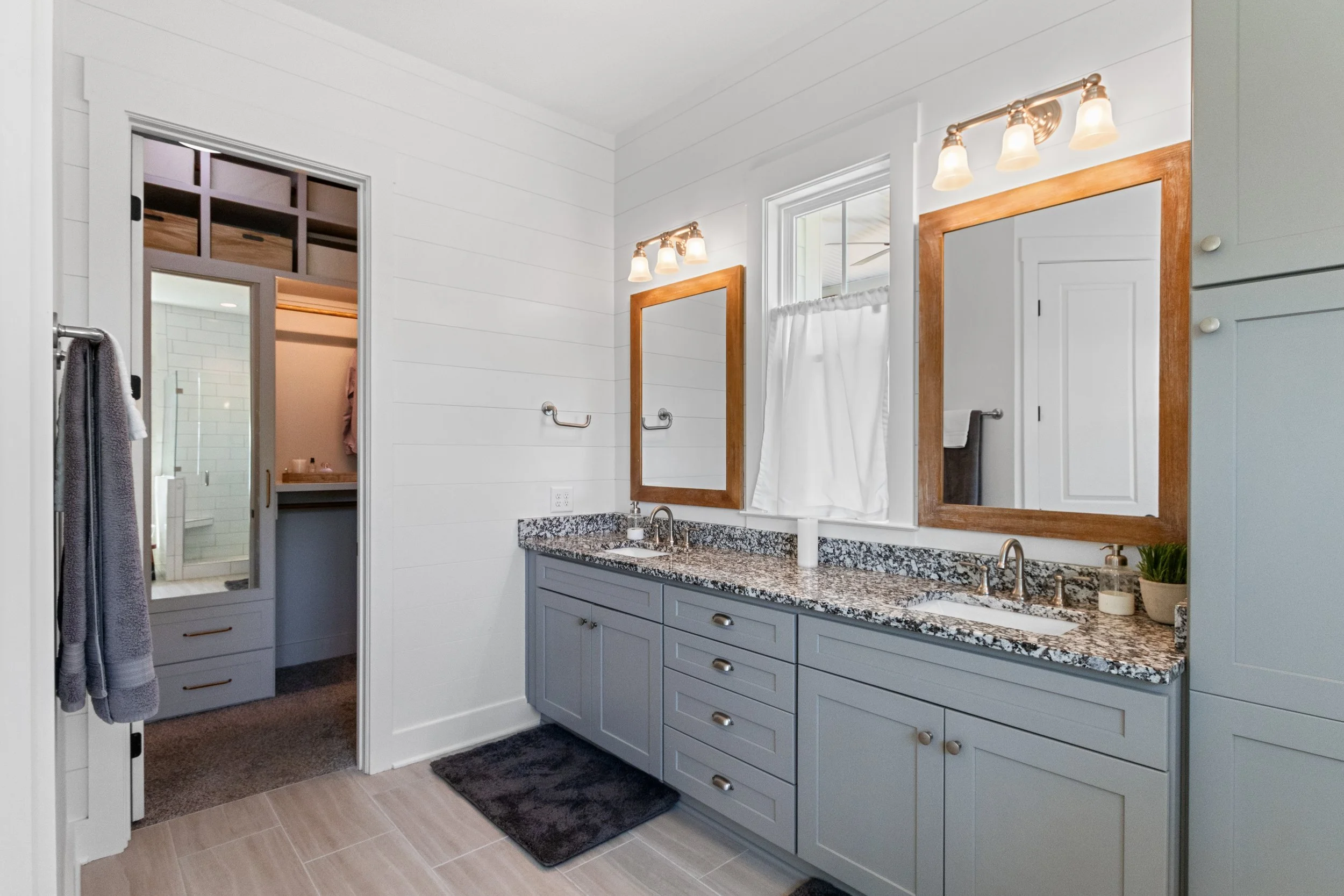 A modern bathroom with a double vanity with gray cabinets, granite countertops, two mirrors with wooden frames, and two sets of three-light vanity fixtures. There's a window with a white curtain, a greenery plant, and a soap dispenser. The bathroom l