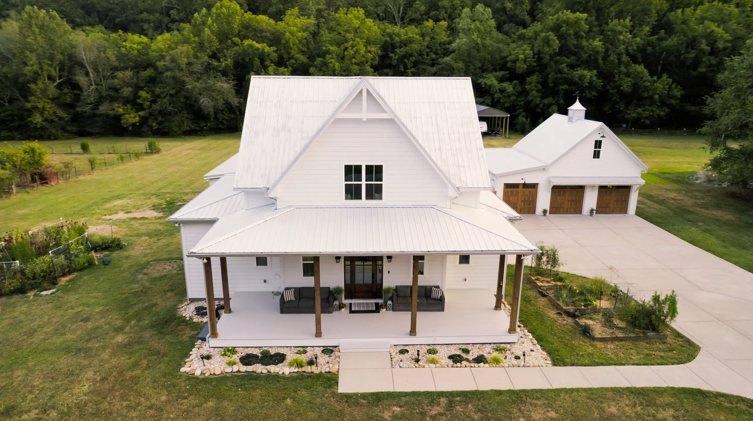 A white farmhouse with a metal roof, front porch with seating, landscaped garden, concrete driveway, and a detached garage, surrounded by greenery and trees.