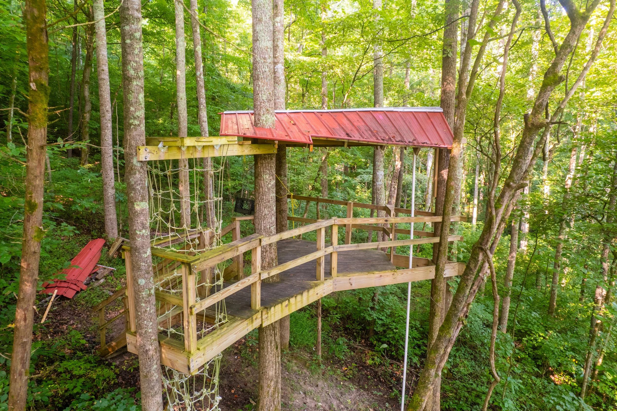 Treehouse with a red metal roof built among tall trees in a lush green forest, featuring a wooden deck and climbing ropes.