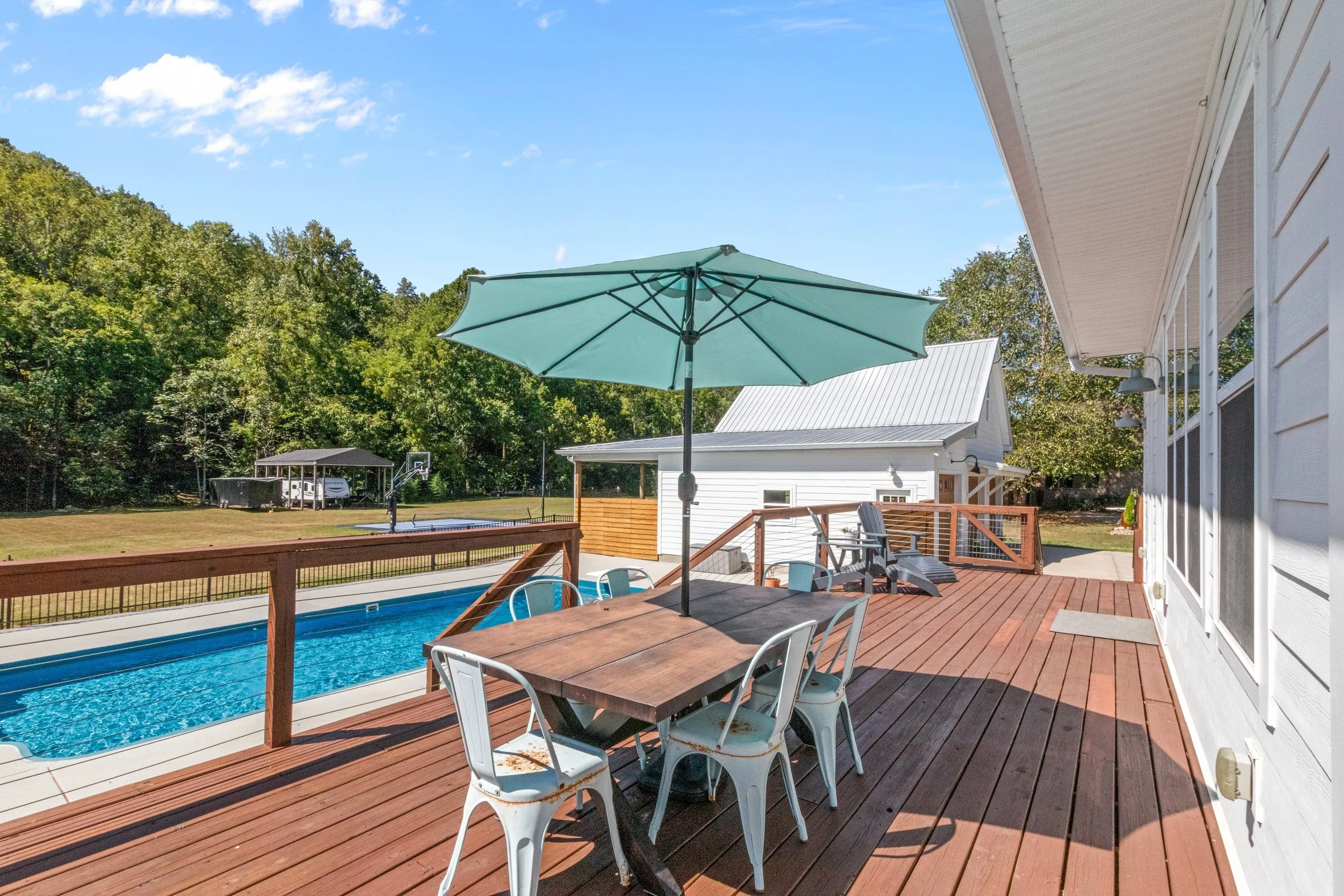 Backyard with a wooden deck, swimming pool, patio table with an umbrella, Adirondack chairs, and a grassy yard with a basketball hoop and shed under a blue sky.