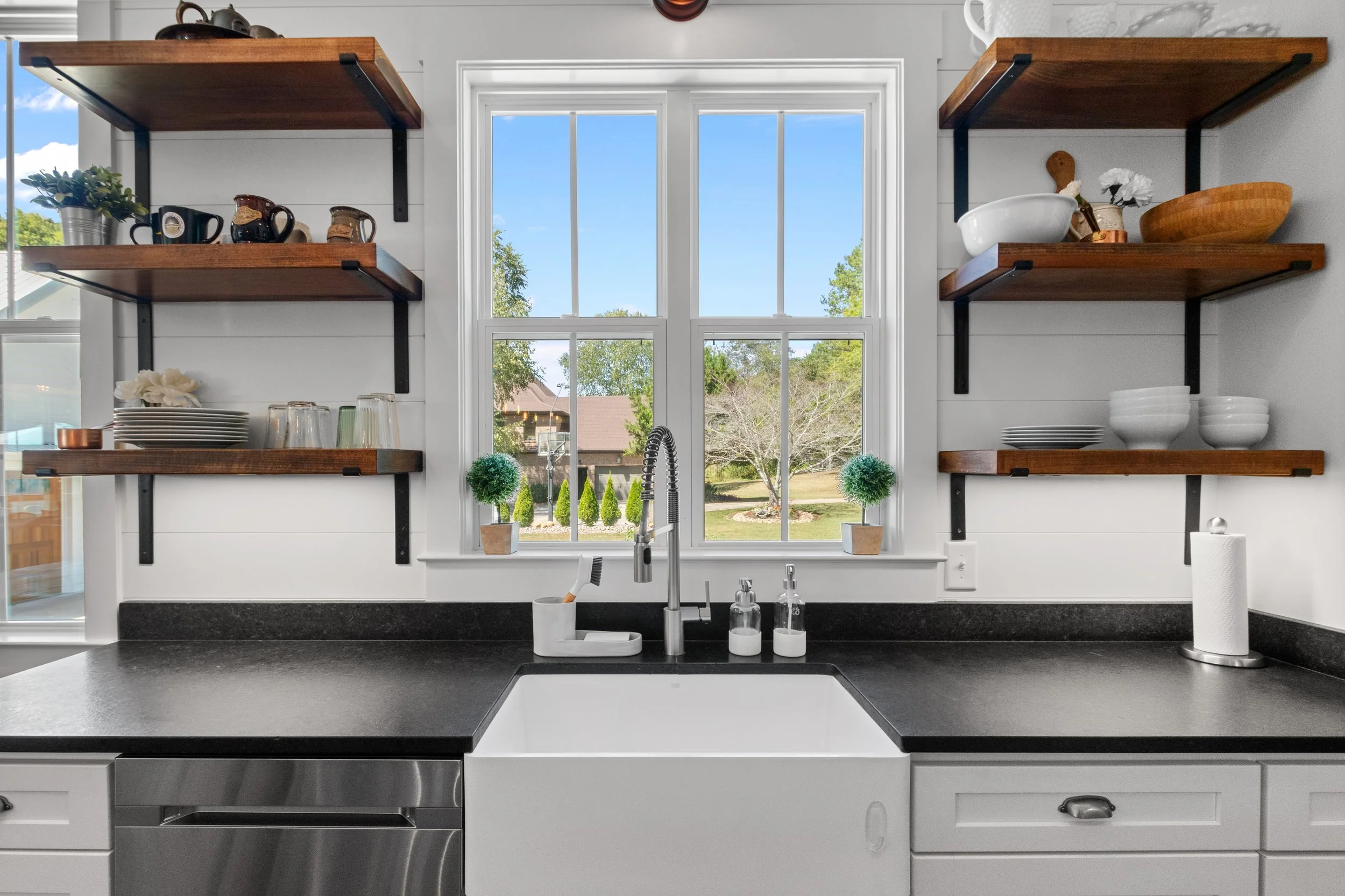 Kitchen sink area with black countertop, white farm sink, and a window showing a green backyard with trees and houses. Open wooden shelves on either side hold dishes, bowls, cups, a plant, and decorative items. There are two small potted plants on th