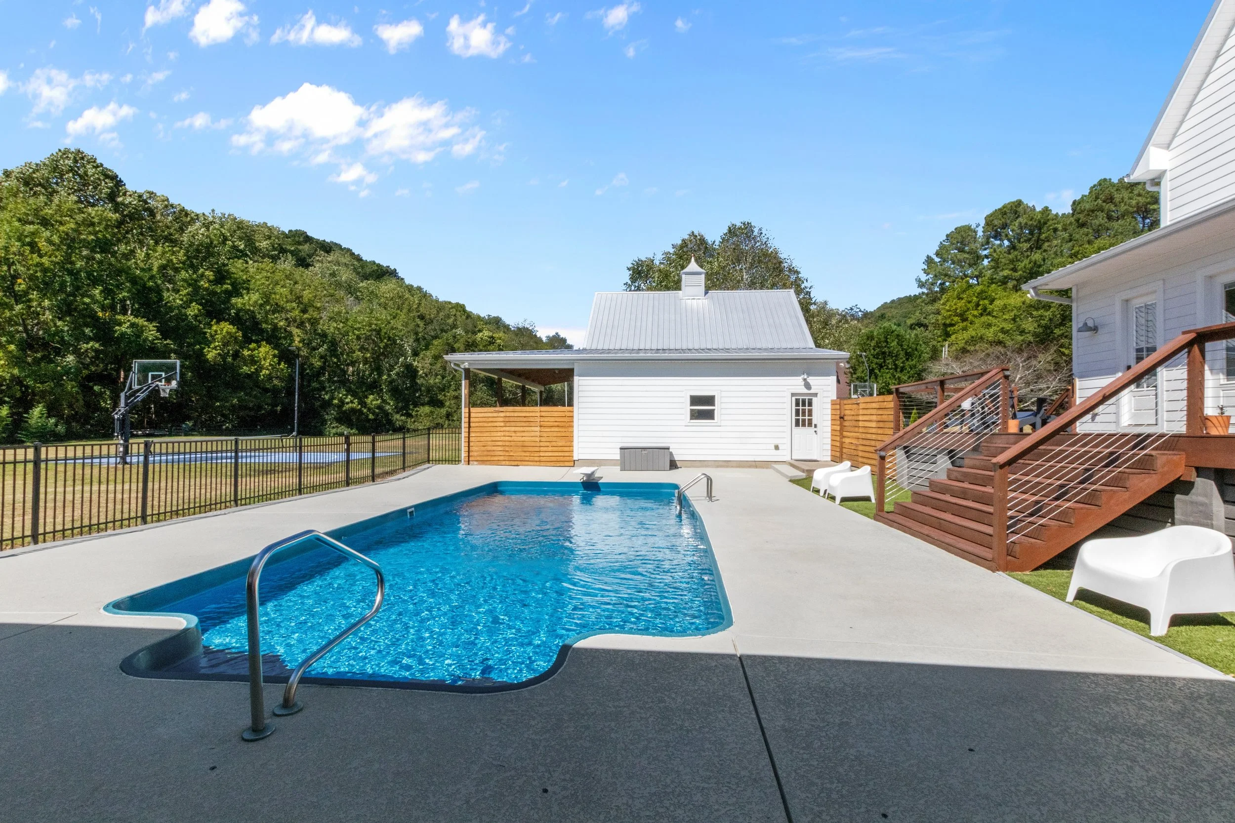 Residential backyard with a swimming pool, a small basketball hoop, a white house, a deck with stairs, outdoor seating, and green trees in the background under a blue sky.