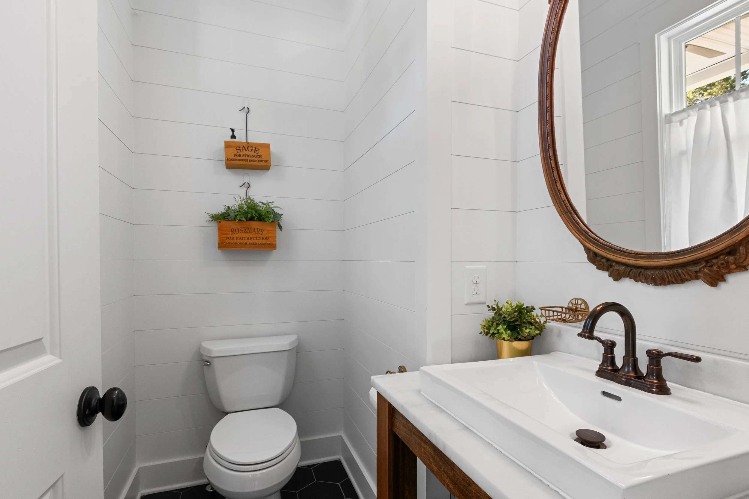 A small bathroom with white shiplap walls, a white toilet, a wooden vanity with a white sink, a bronze faucet, a round wooden framed mirror, a small potted plant in a gold pot, and a window with white curtains. There are two wooden hanging planters w