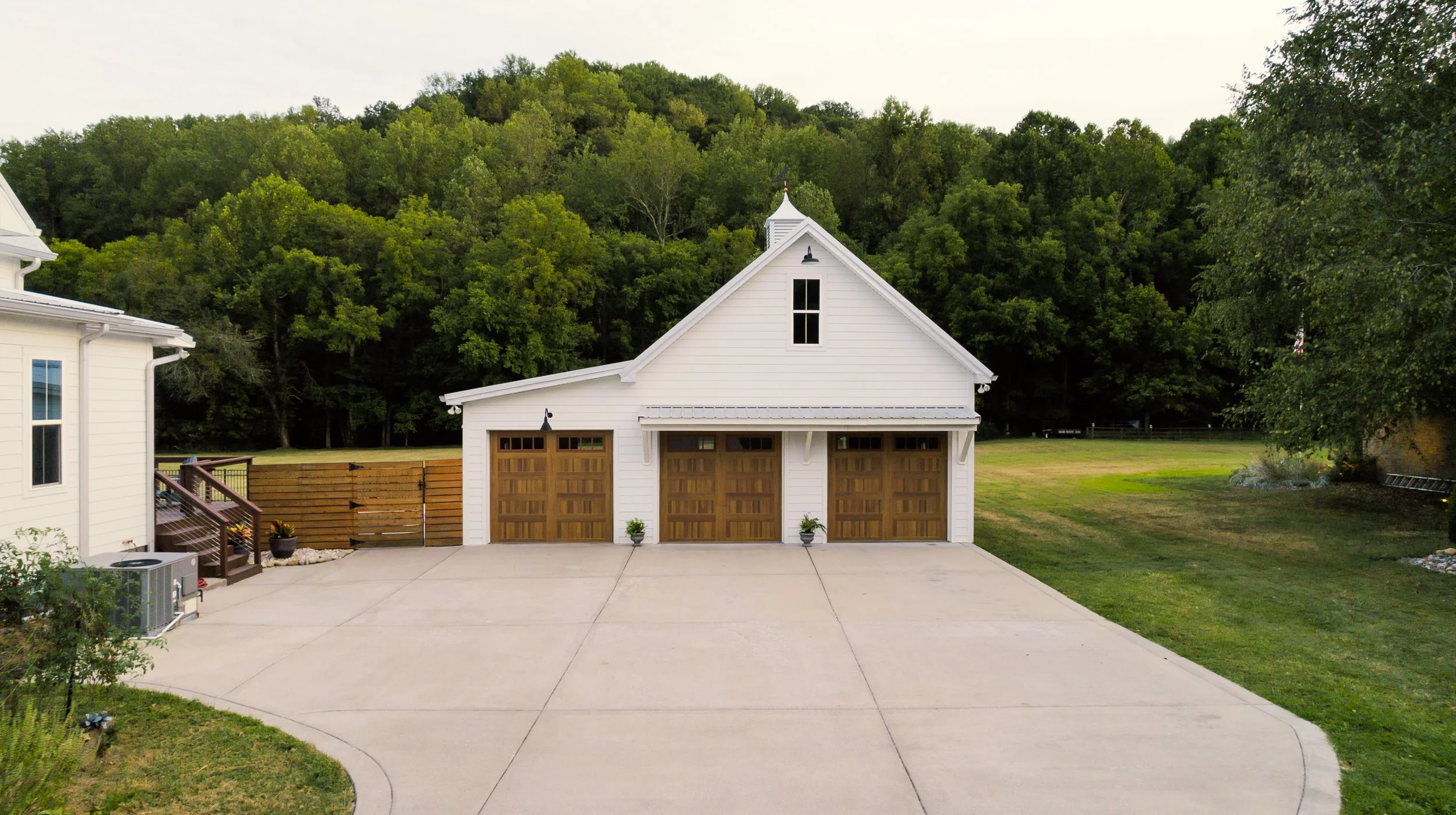 A white garage building with three wooden garage doors, a concrete driveway in front, surrounded by a grassy lawn and trees in the background.