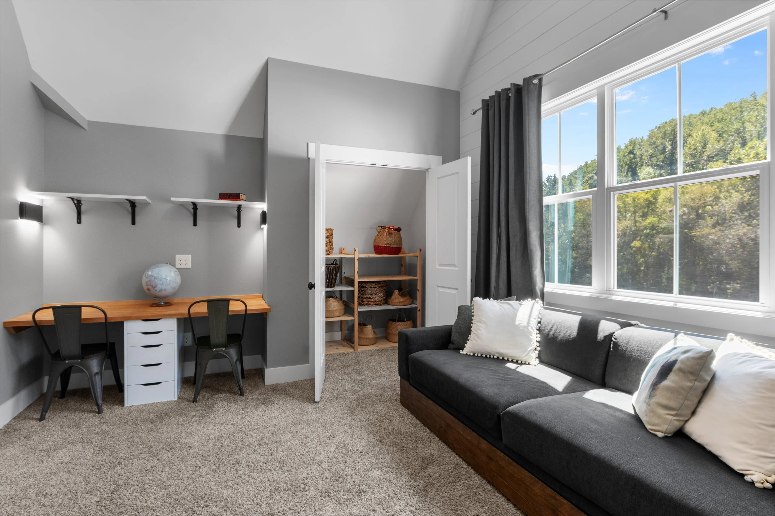 A room with gray walls, a desk with two black chairs, a globe, wall shelves, a dark gray sofa with white pillows, large window with dark curtains, and a shelf with baskets inside a closet.