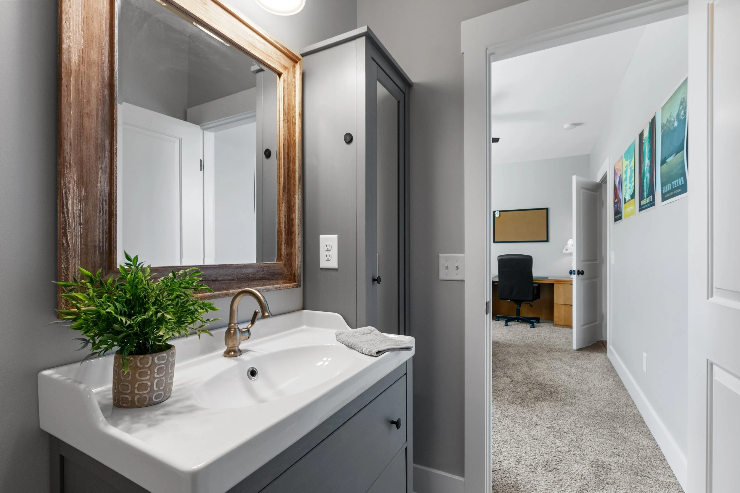 Bathroom with a gray vanity and white sink, decorated with a potted green plant and a gray hand towel, mirror above the sink, doorway leading to a home office room with a desk, chair, and colorful posters on the wall.