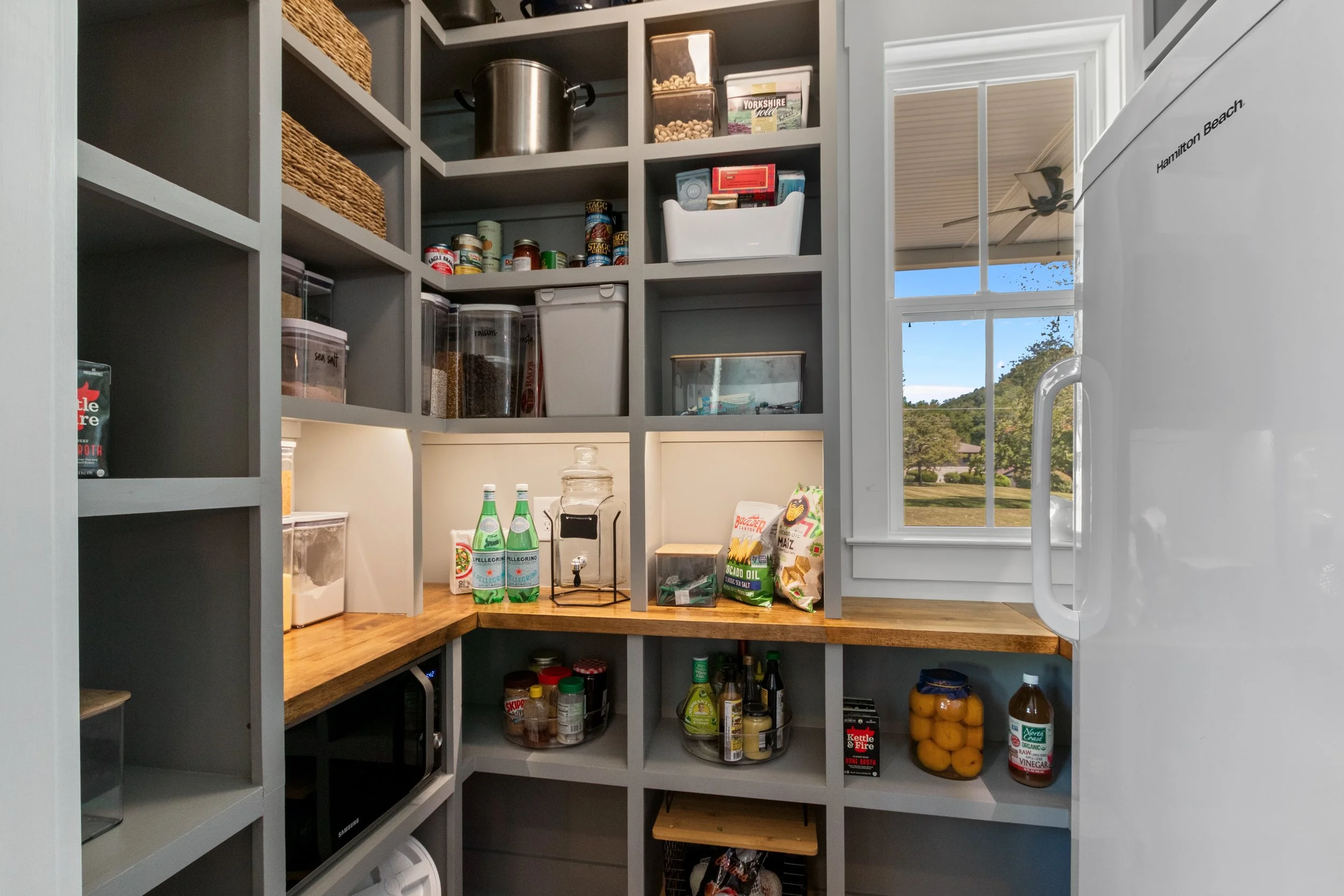 Pantry with gray shelving, containing jars, canned goods, bottles, a microwave, and a countertop with bottled water, snacks, and canned food. A window shows an outdoor scene with a lawn and trees.