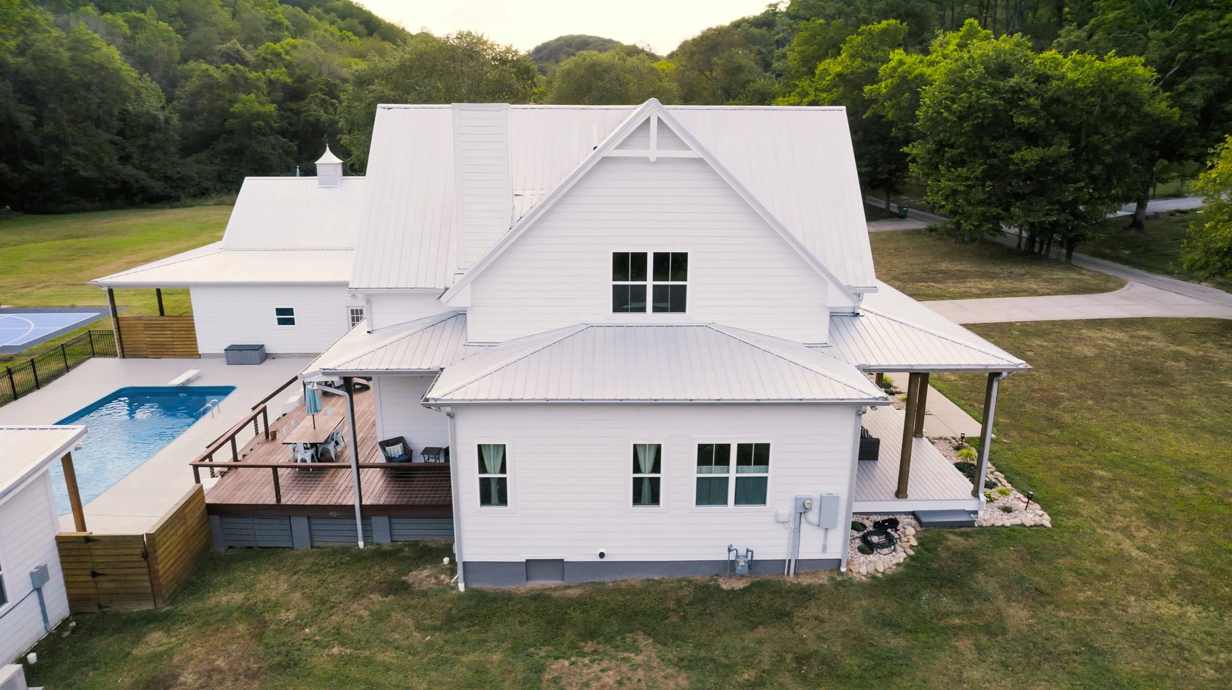 A white house with a metal roof, a deck with outdoor furniture, a swimming pool, surrounded by green grass and trees, with a wooded area in the background.