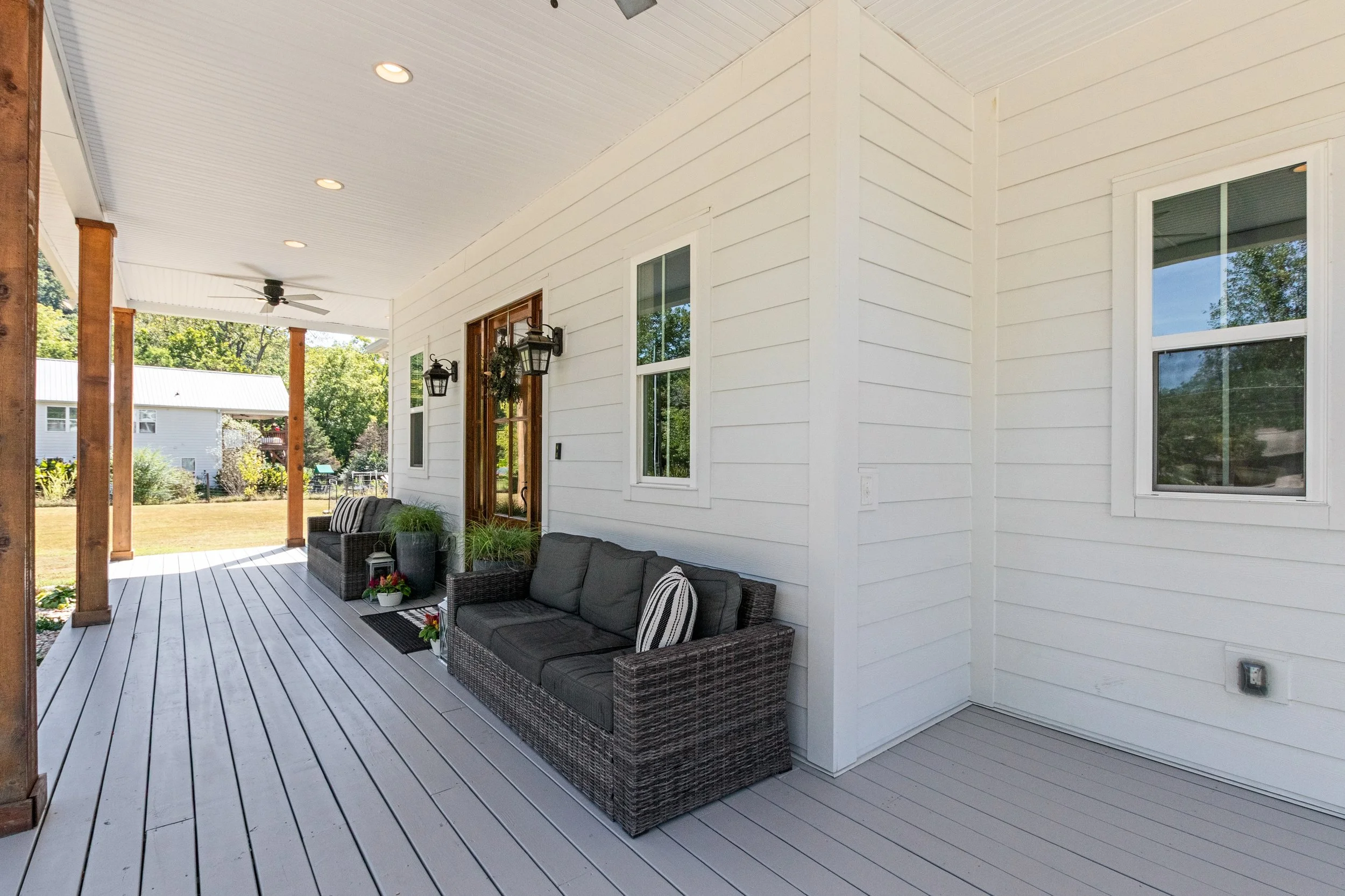 Front porch of a house with white siding, two dark wicker sofas with black cushions and striped pillows, potted plants, wall-mounted lanterns, a glass door with a wreath, and windows, with a view of a backyard with trees and neighboring houses.