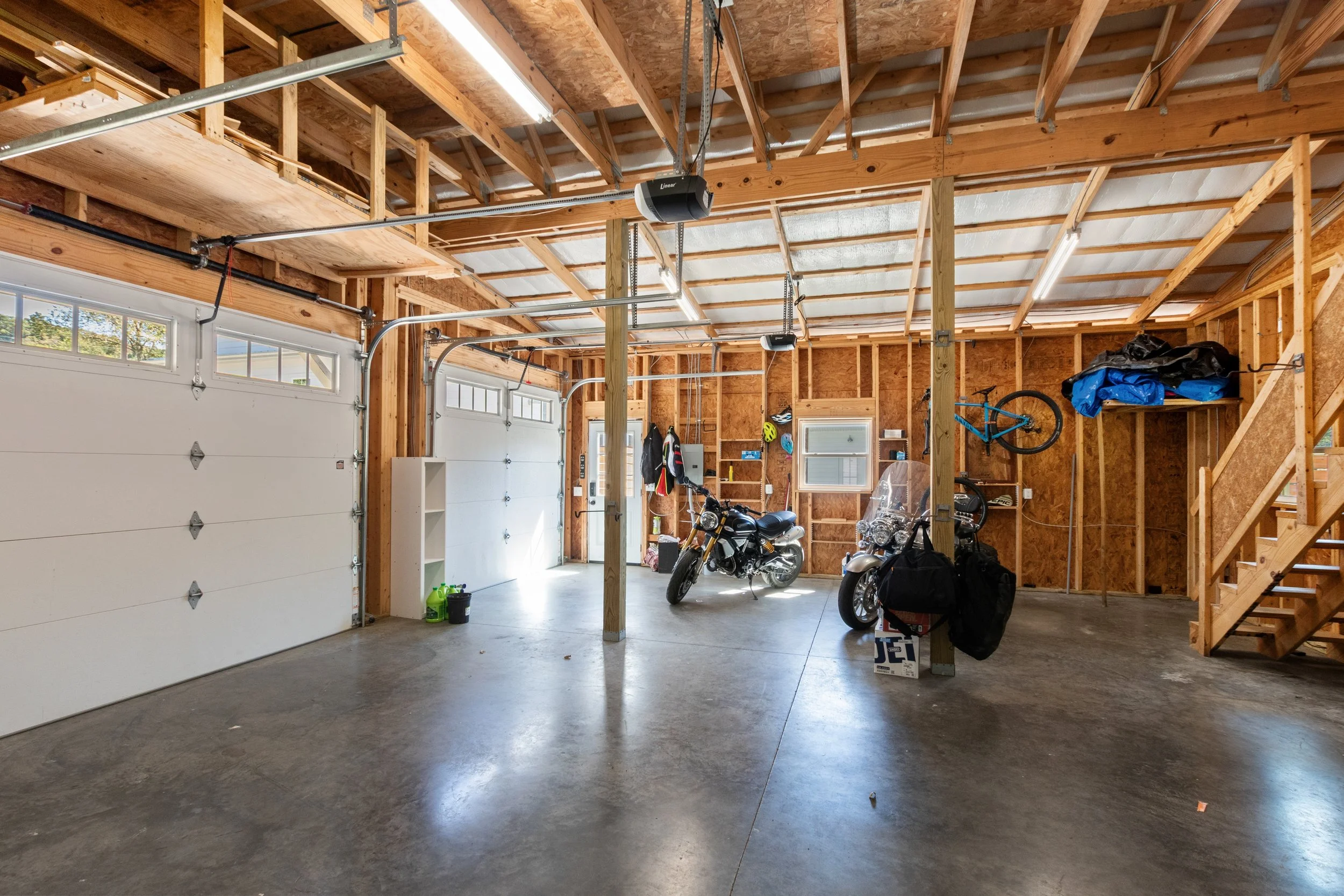 A spacious two-car garage with unfinished wooden walls and ceiling, concrete floor, two white garage doors, motorcycles, biking gear, and storage shelves.