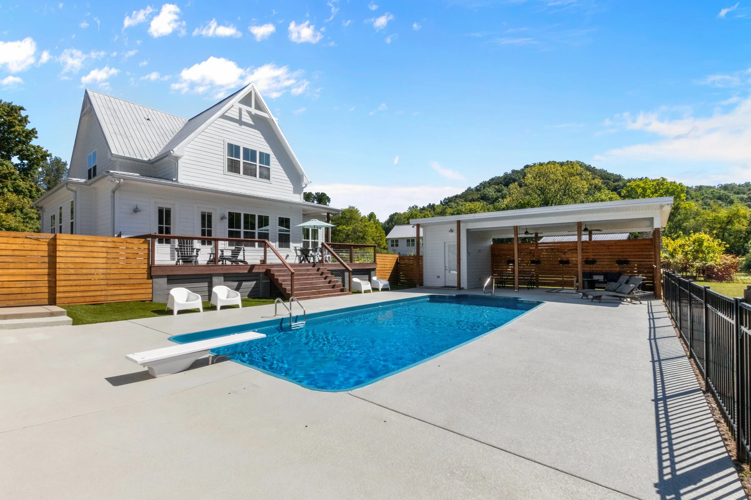 A backyard with a white house, a swimming pool, and a covered outdoor seating area with shade and outdoor furniture, surrounded by a black fence and wooden privacy fence, with trees and hills in the background.