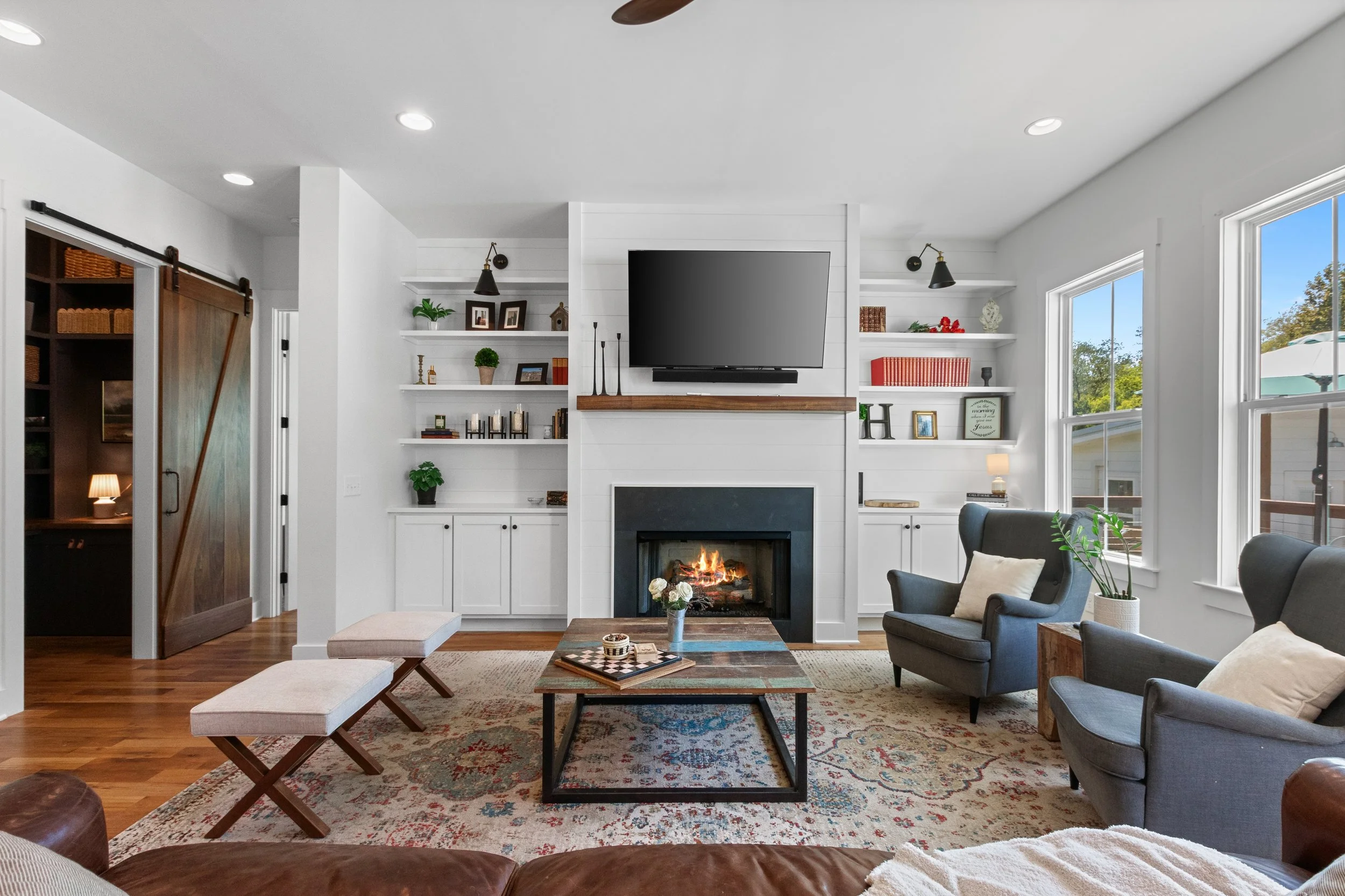 Living room with a fireplace, wall-mounted TV, built-in bookshelves, gray armchairs, a coffee table, windows, and decorative items.