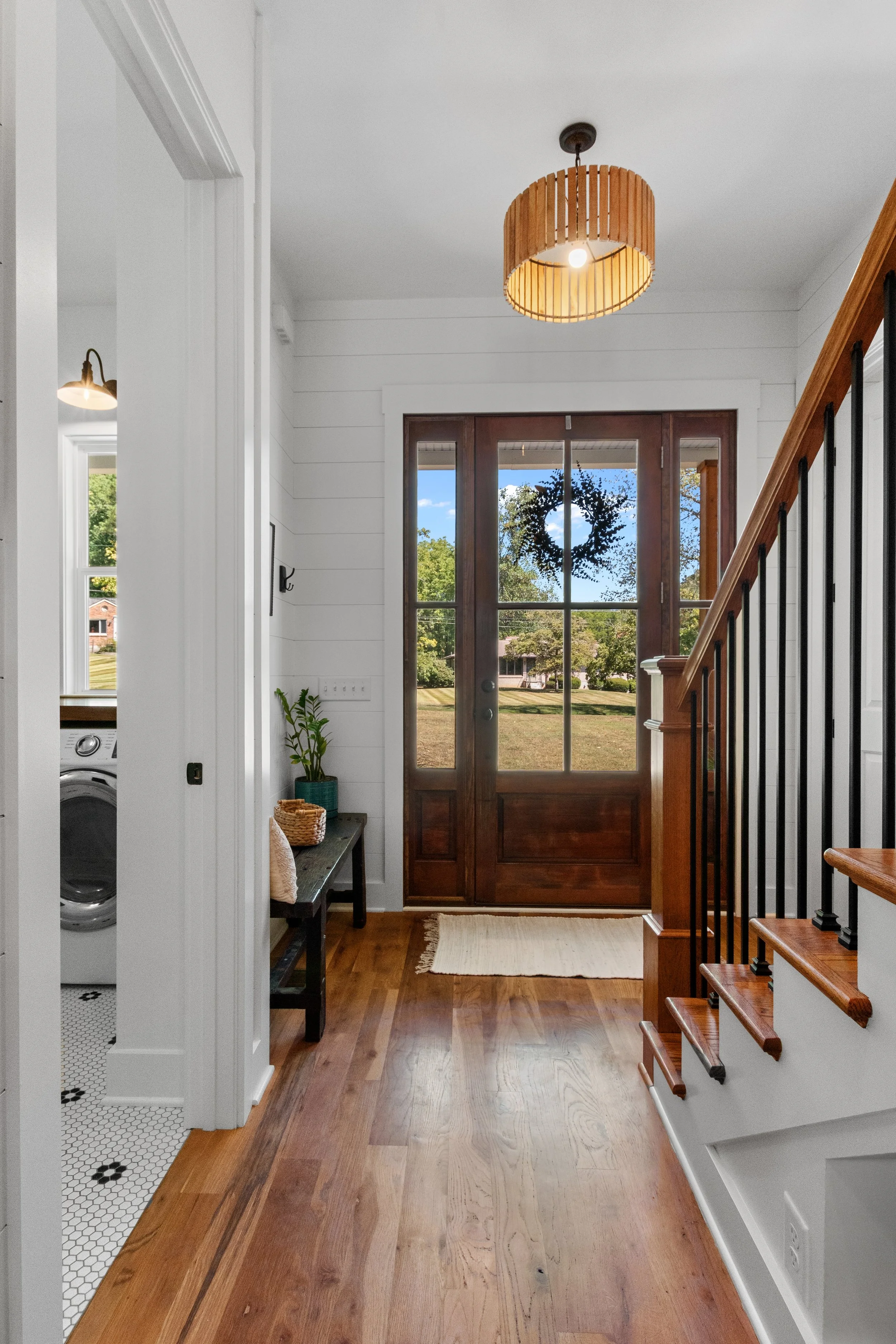 Entryway of a house with a wooden front door and large glass panels, wooden stairs with black balusters, pendant light with a wooden shade, small black bench with a potted plant, woven basket, and a rug inside. A partial view of a laundry area with a