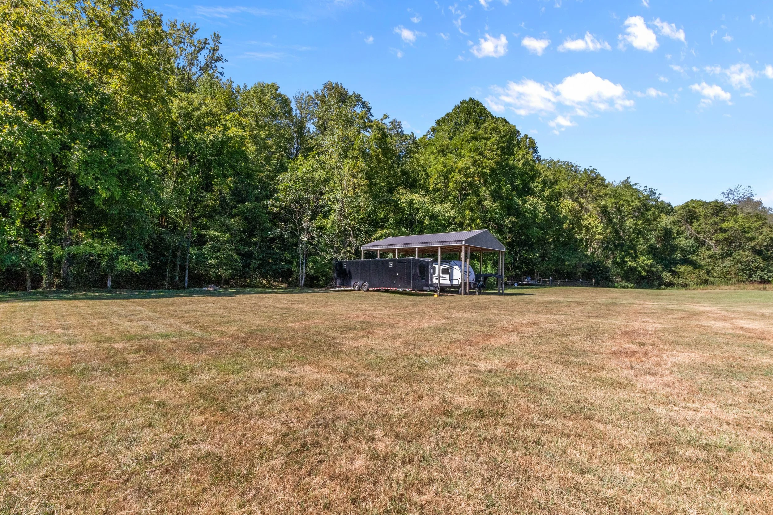 A grassy field with a covered area housing a mobile home or RV and a black trailer, surrounded by trees under a partly cloudy sky.