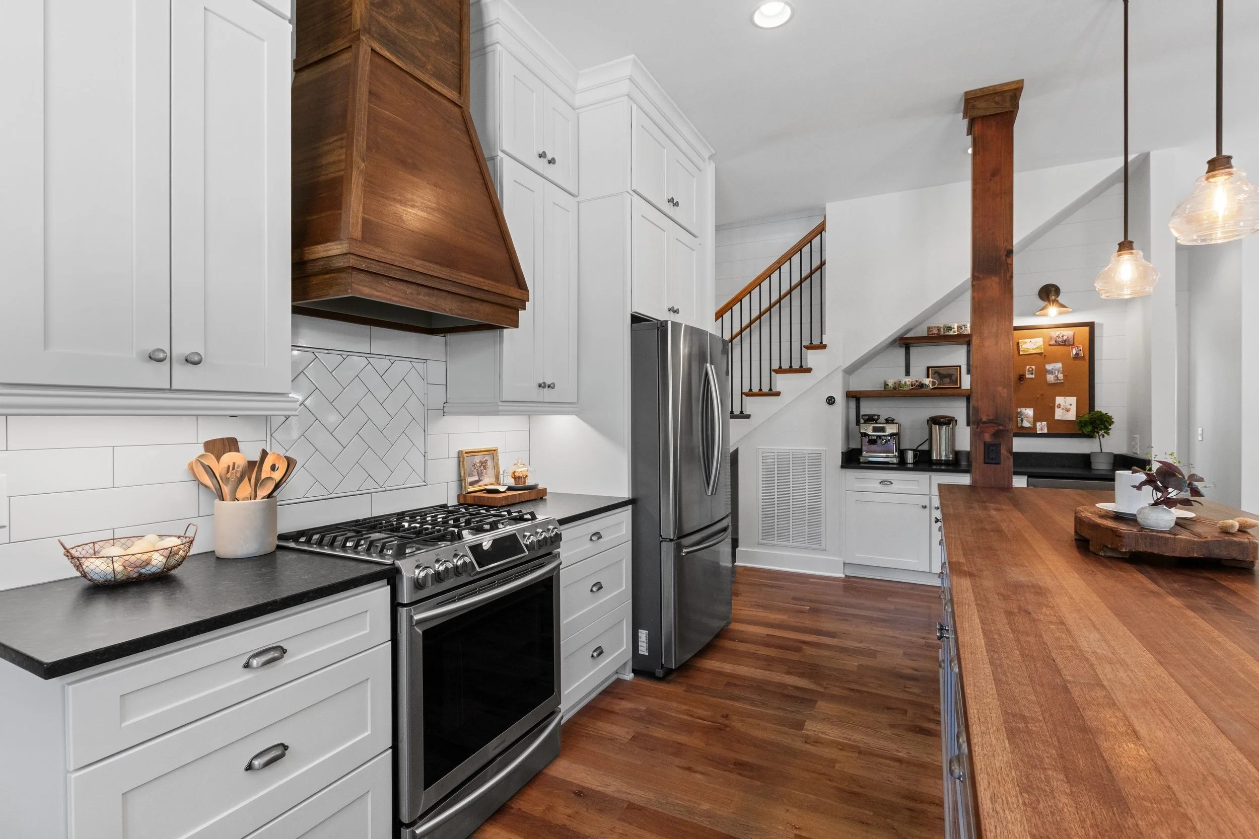 A modern kitchen with white cabinetry, black countertops, a stainless steel refrigerator, and hardwood flooring. There is a wooden range hood above a stove, decorative items on the counters, and a staircase in the background with a small desk area ne
