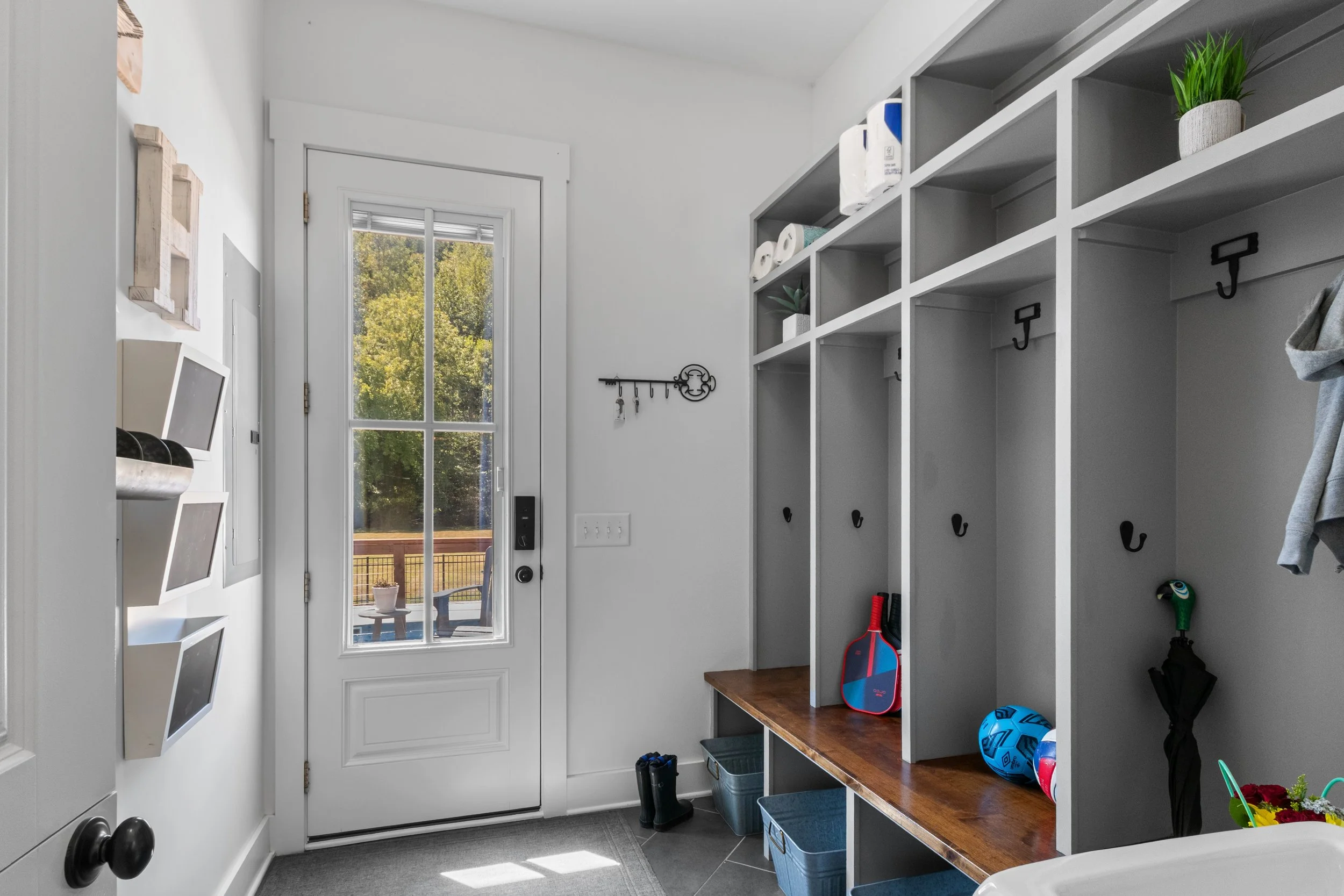 Interior of a mudroom with a white door, gray cubbies, hooks for hanging, and various items like toys, shoes, and plants.