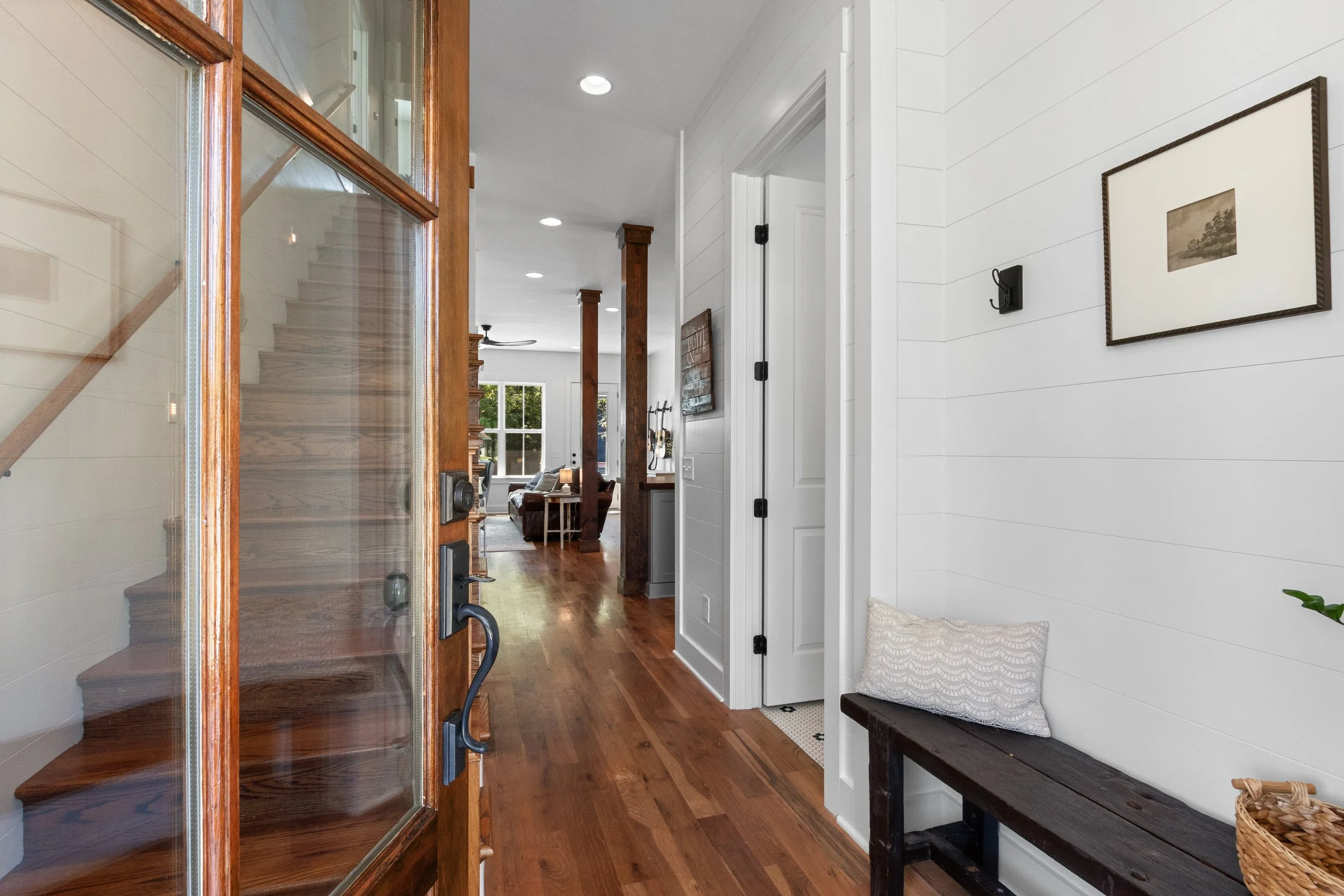 Interior view of a home entryway with a wooden door, hardwood floors, white shiplap walls, and a bench with a pillow on it.