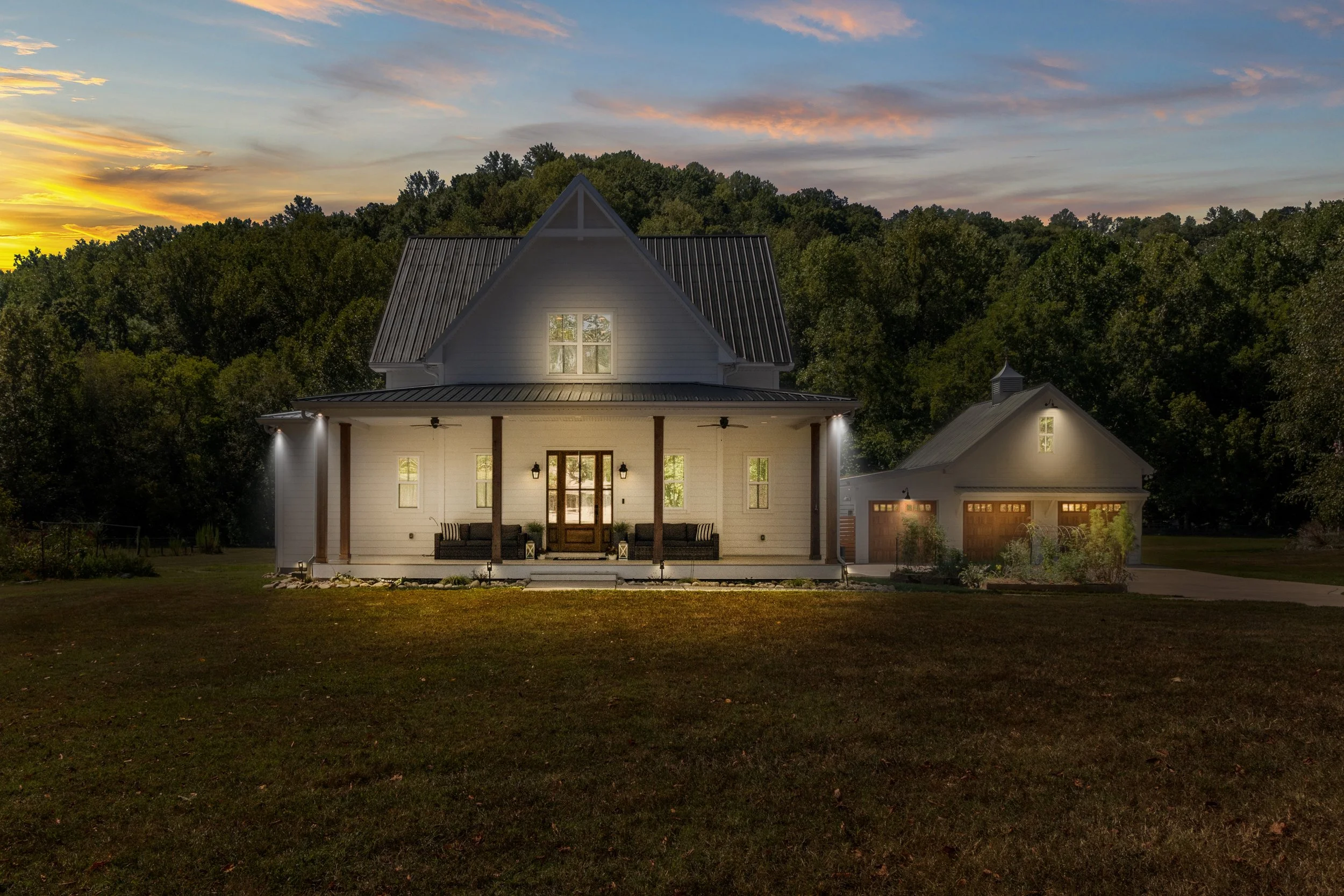 A modern farmhouse-style house with a porch, illuminated by outdoor lights at dusk, surrounded by trees and a grassy lawn.