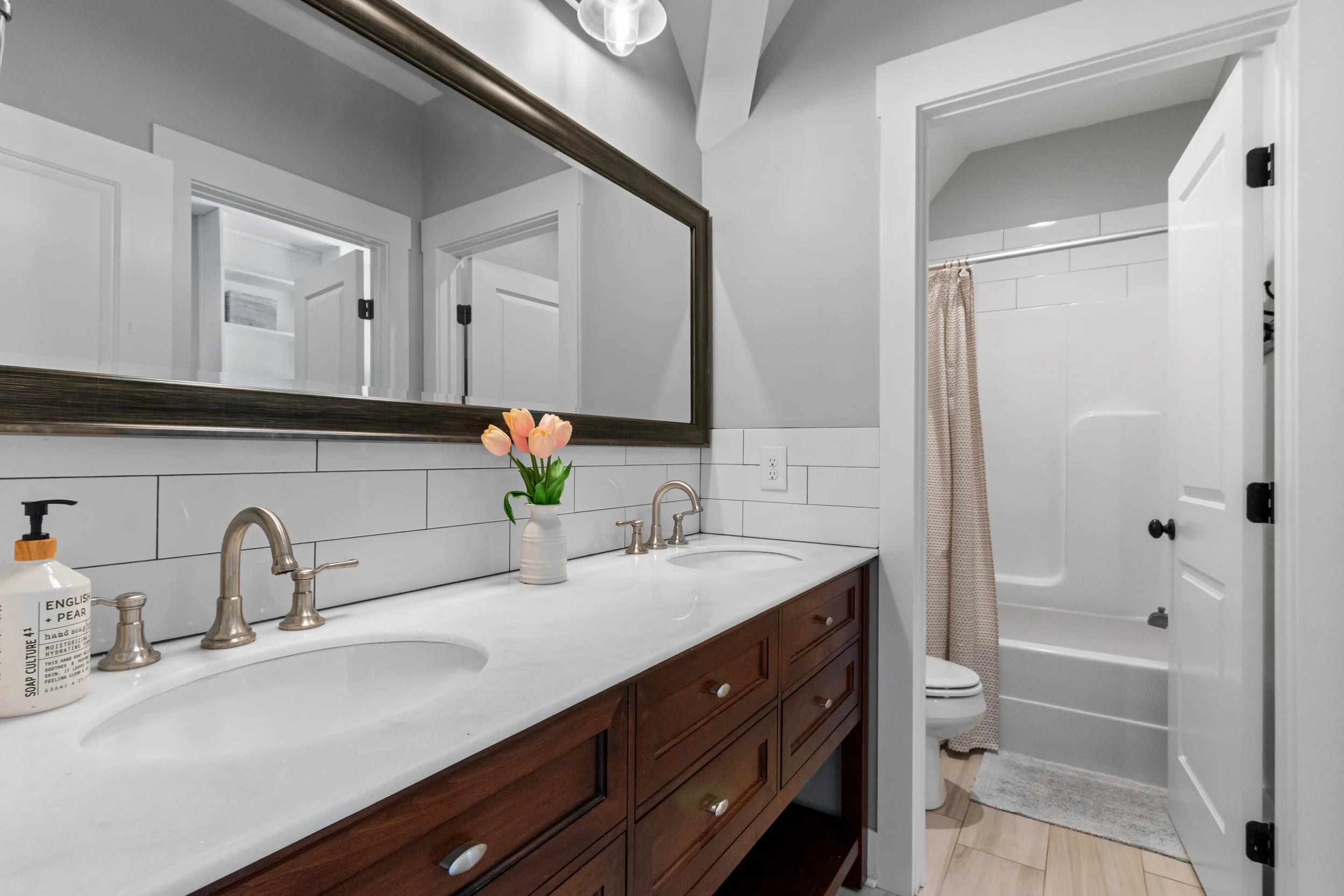 Bathroom with a double vanity, large mirror, faucet fixtures, a vase with pink tulips, and a shower/bathtub with a curtain in a small room.