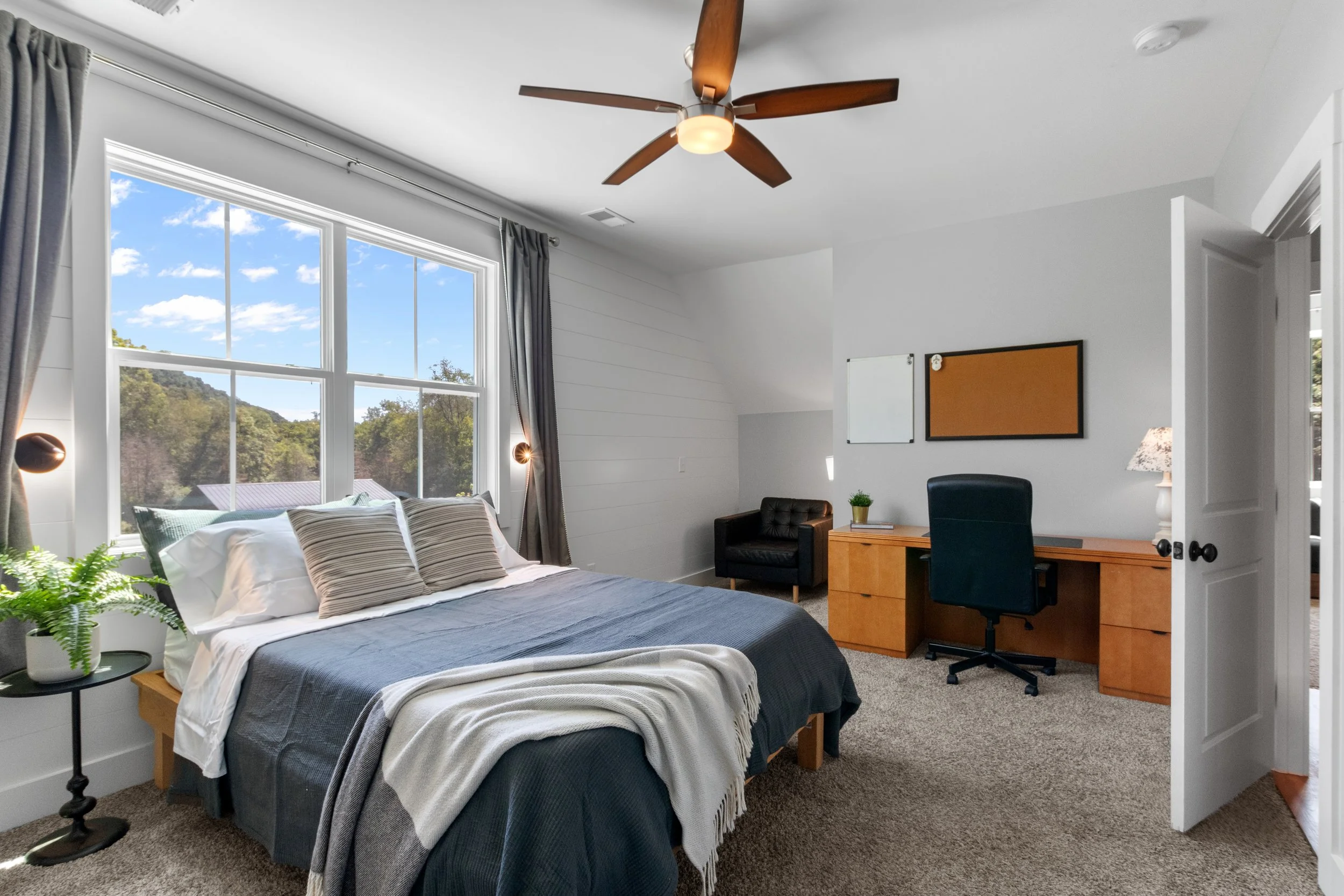 Bedroom with large window, bed with pillows and blanket, desk with chair, black armchair, ceiling fan, wall-mounted whiteboard and corkboard, side table with plant, carpeted floor.