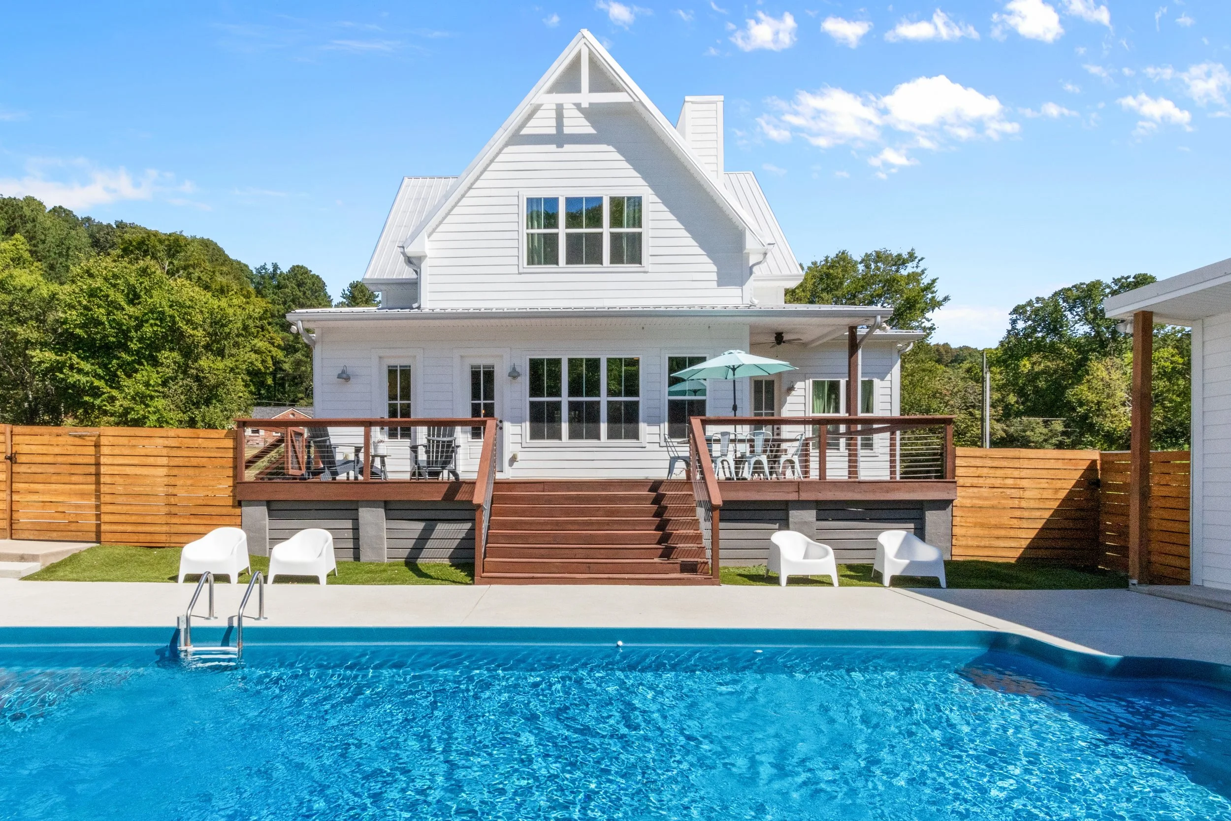 A white two-story house with a deck, outdoor seating, and a swimming pool in the foreground, surrounded by a wooden fence and green trees under a blue sky.