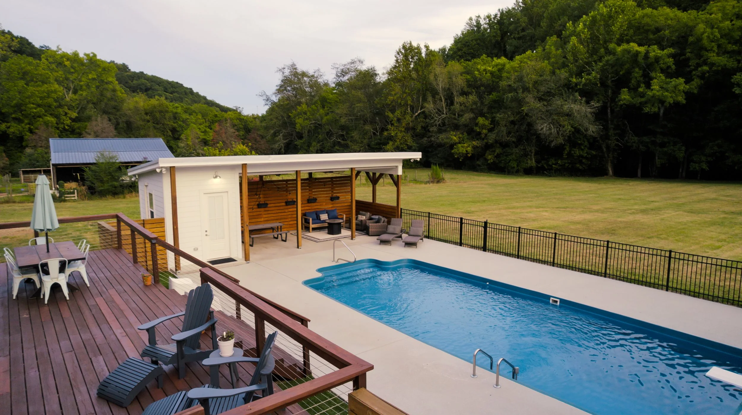 View of a backyard with a swimming pool, sitting area, wooden deck, and surrounding green field and trees.
