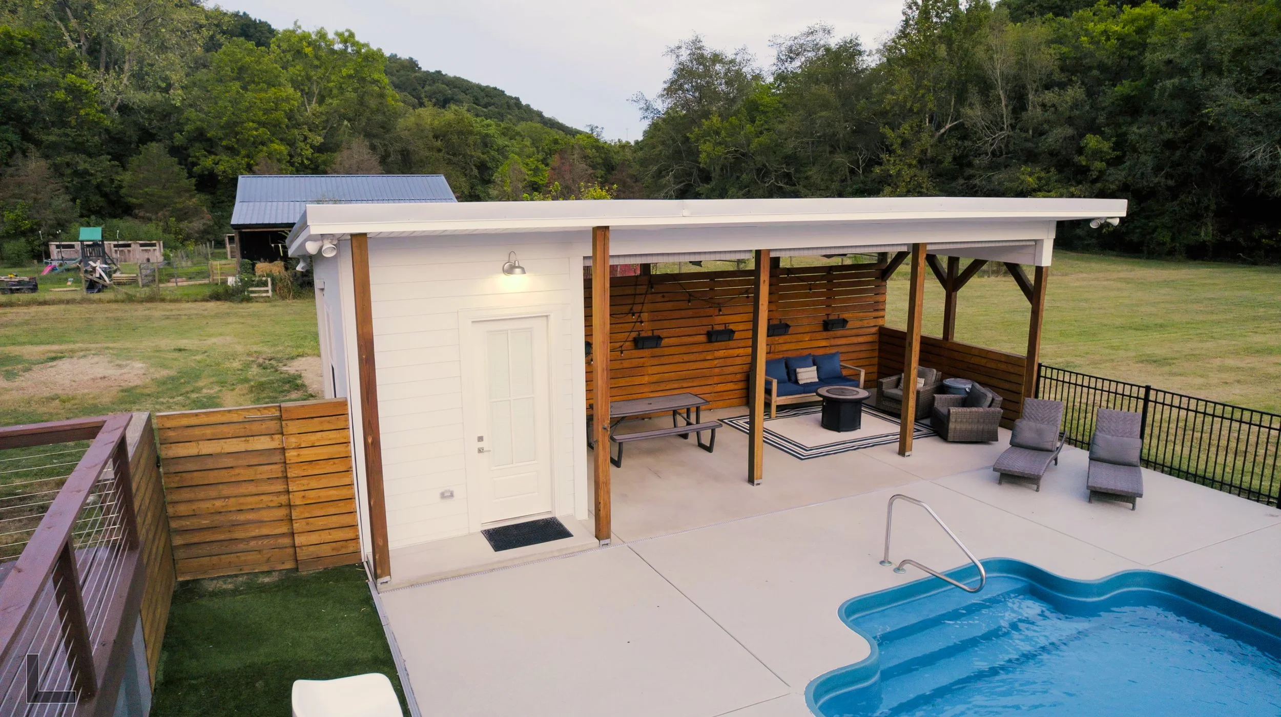 Residential backyard with a swimming pool, patio furniture, a small building, and a fenced grassy yard, with trees in the background.