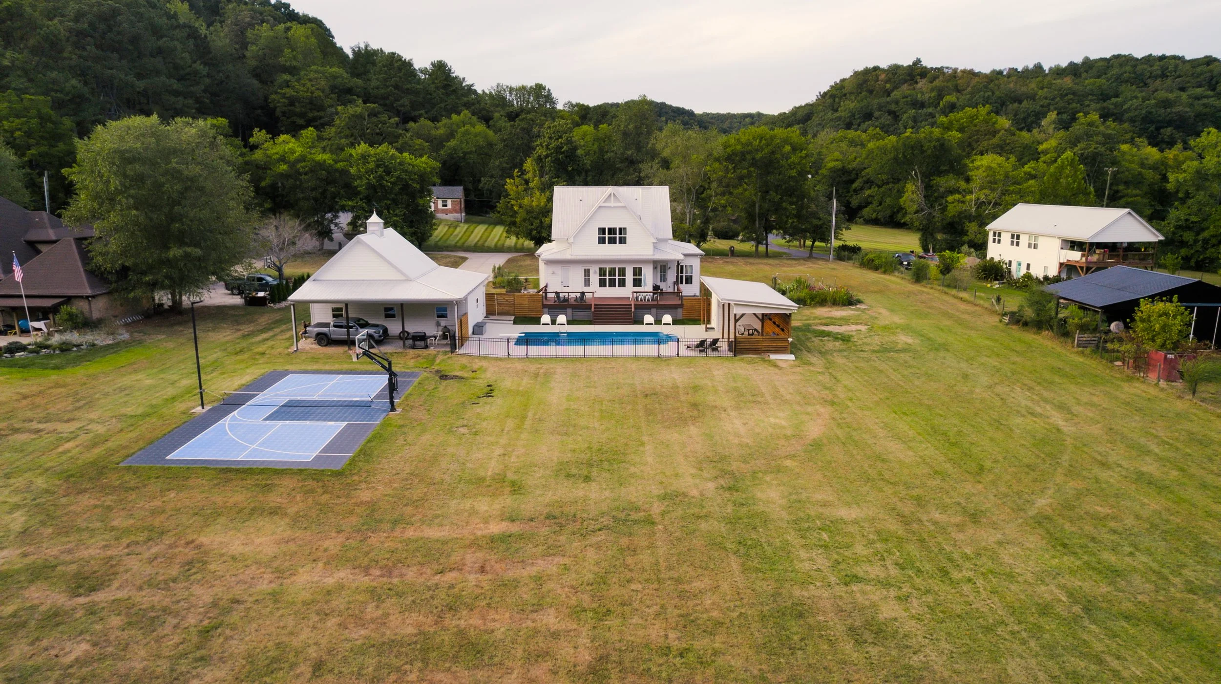Aerial view of a large backyard with a white house in the center, a swimming pool, a basketball court, and surrounding trees and neighboring houses.
