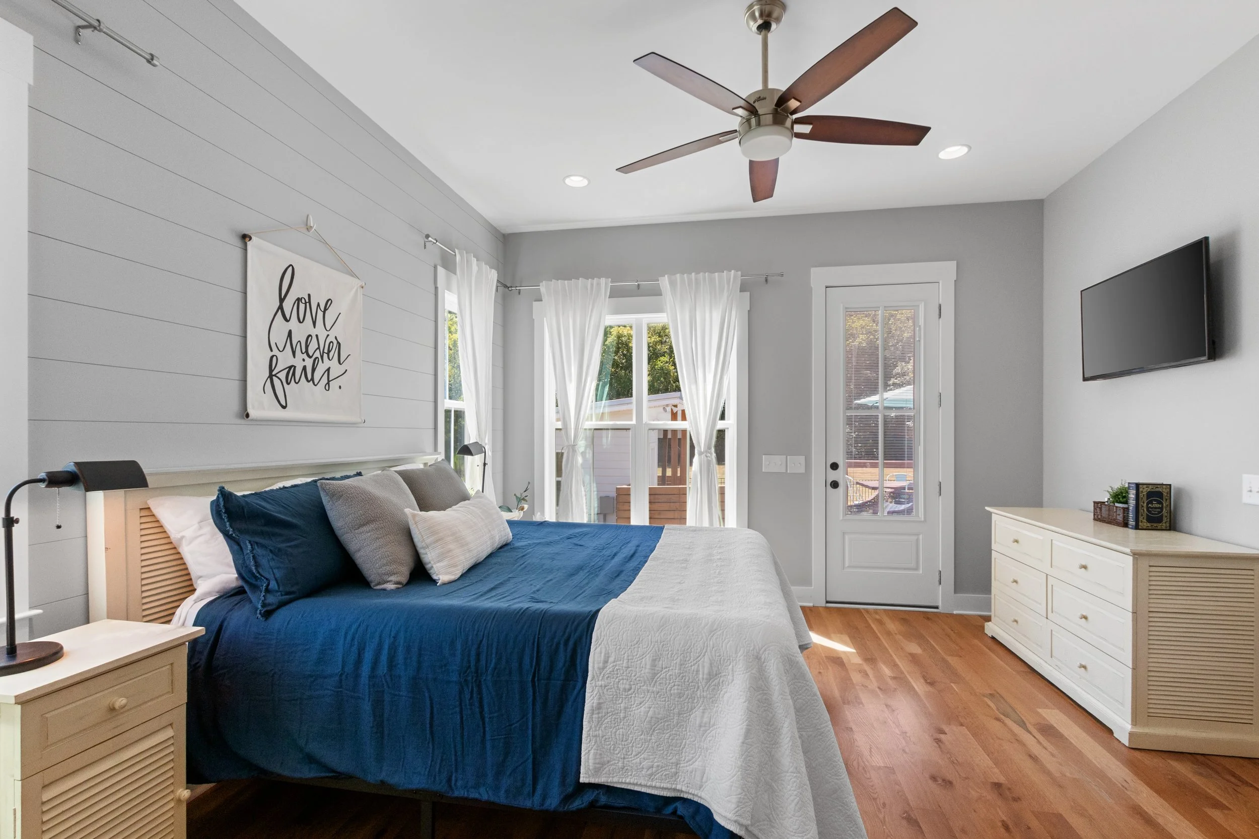 Bright bedroom with white walls, a ceiling fan, a bed with navy blue and white bedding, decorative pillows, a window with white curtains, and a white dresser with small items on top.