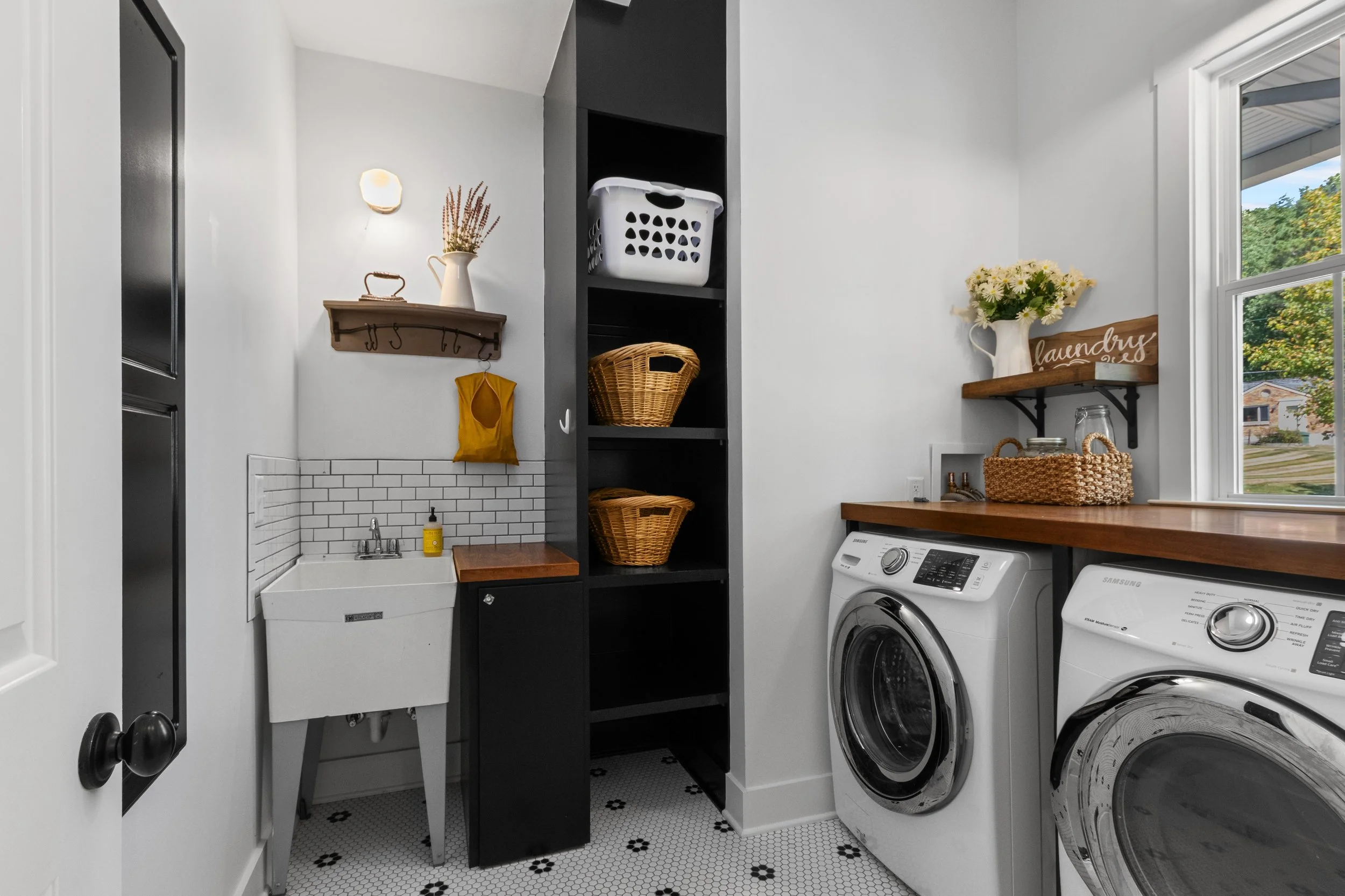A laundry room with a white utility sink, black shelving unit, front-loading washing machine and dryer, wooden countertops, woven baskets, and decorative shelves with flowers and laundry signs, natural light coming through a window.