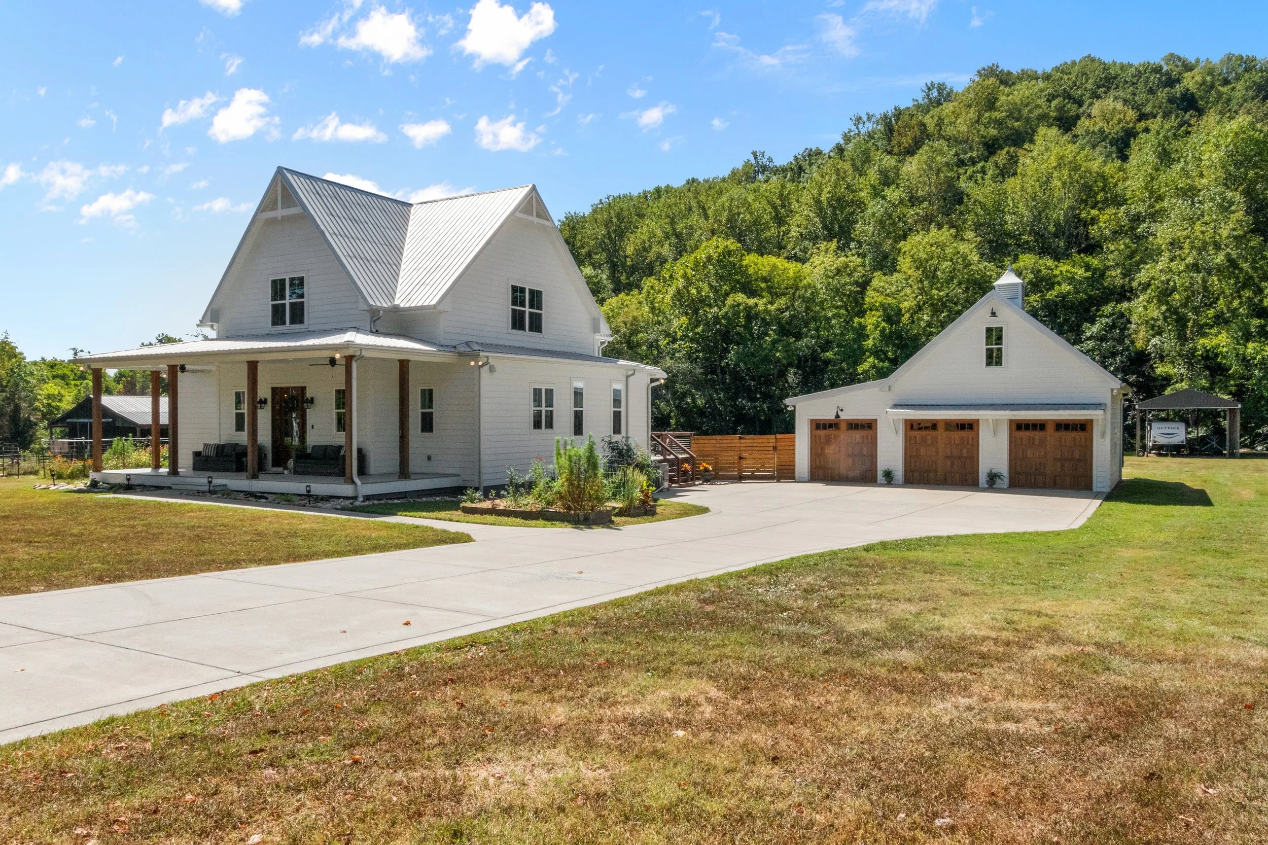 A white two-story house with a covered porch and wooden columns, a detached garage with three wooden doors, a concrete driveway, and a backyard with a green hill and trees.