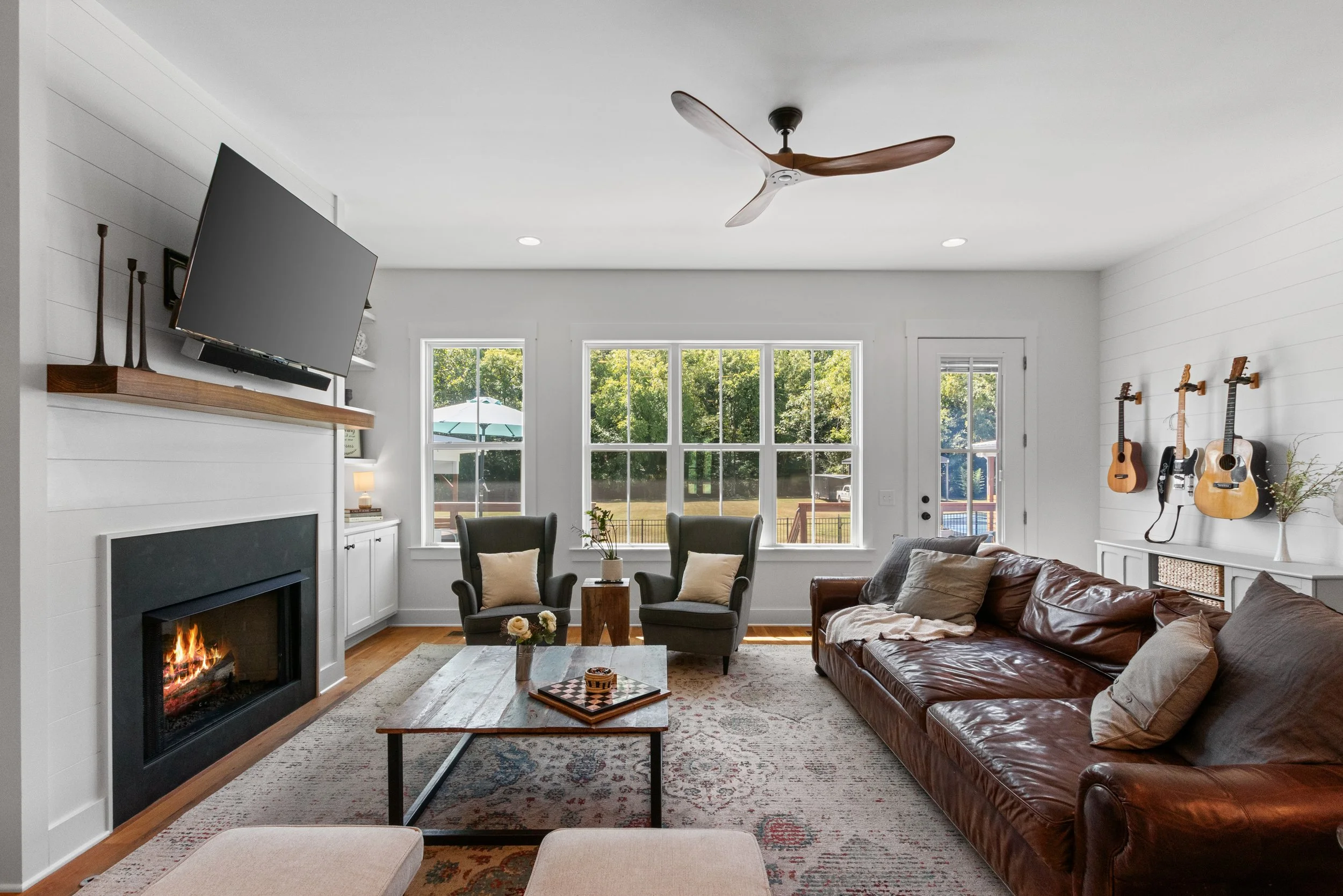 Living room with a fireplace, a mounted TV, two armchairs, a leather sofa, a coffee table, a rug, and guitars on the wall near a windowed door.