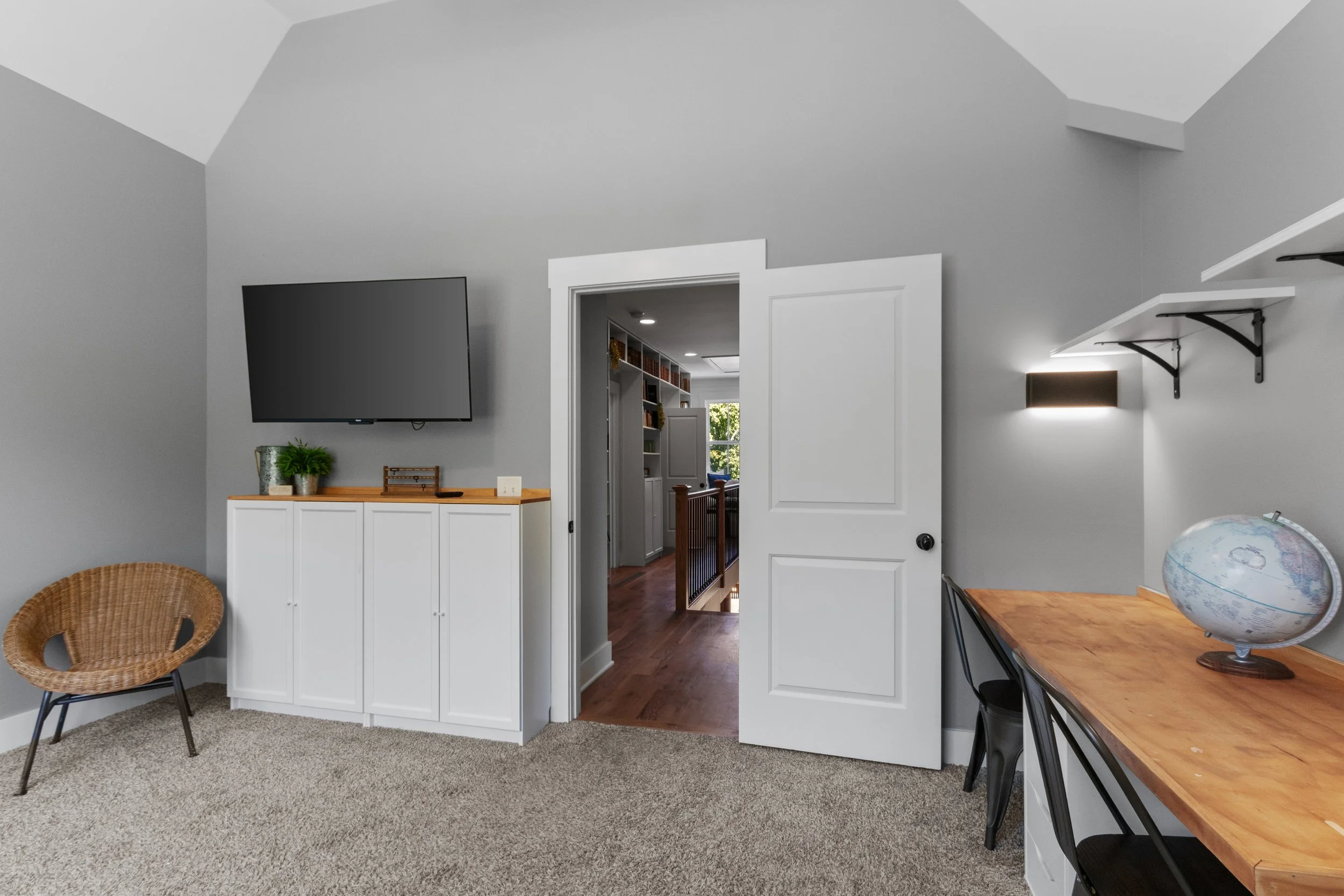 Room with gray walls, a flat-screen TV on a wall, a wicker chair, a white cabinet with plants on top, an open white door leading to a hallway, a wooden desk with a globe, and a black shelf with white brackets.