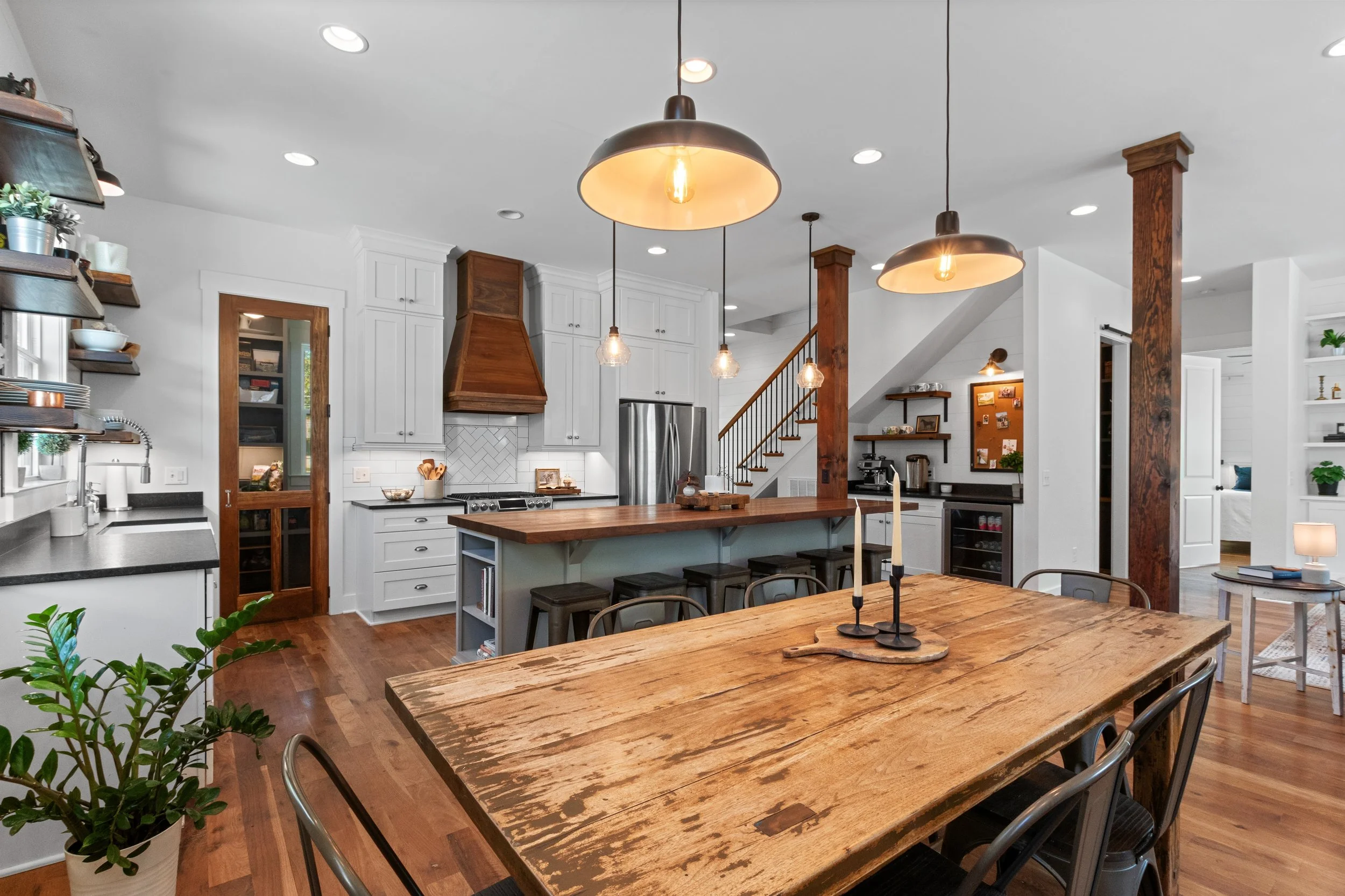 A spacious kitchen with white cabinets, wooden accents, a large wooden dining table, pendant lights, and a staircase in the background.