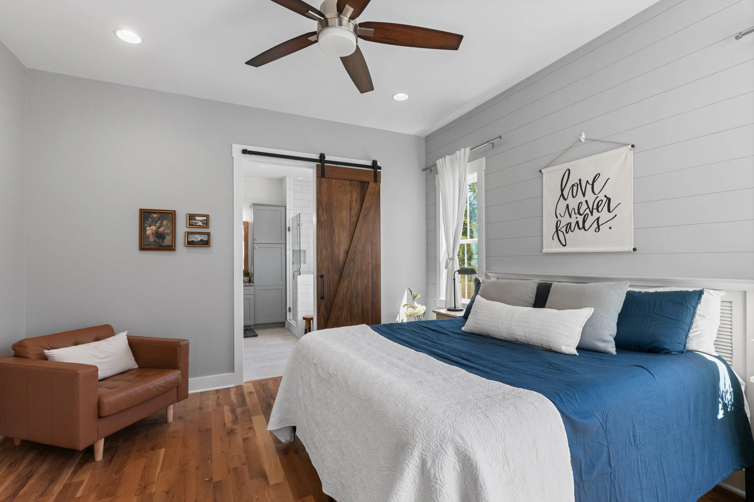 A cozy bedroom with a large bed covered in a white and navy blue quilt, a small leather couch with a white pillow, a ceiling fan, a window with white curtains, and a wall hanging that reads 'love never fails.' There are three small framed pictures on