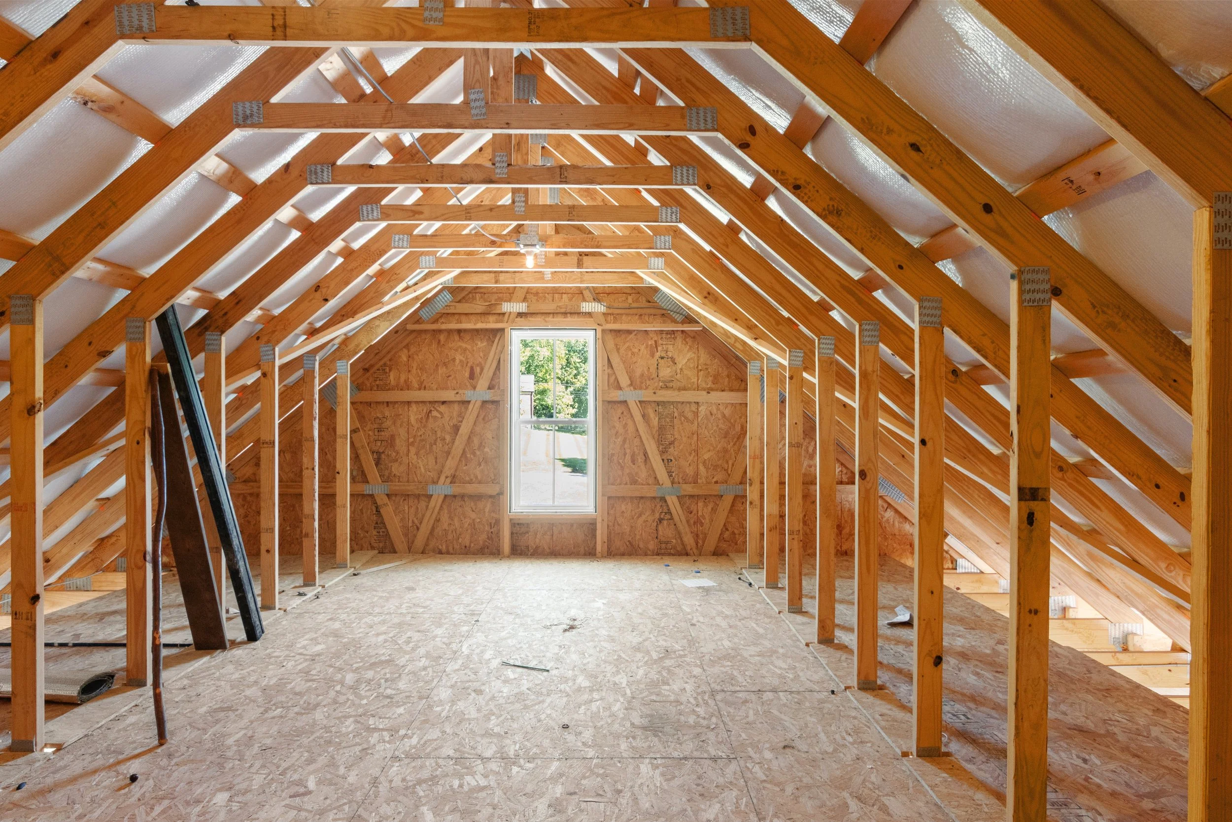 Unfinished attic with exposed wooden framing and plywood floors, featuring a small window at the far end allowing natural light to enter.