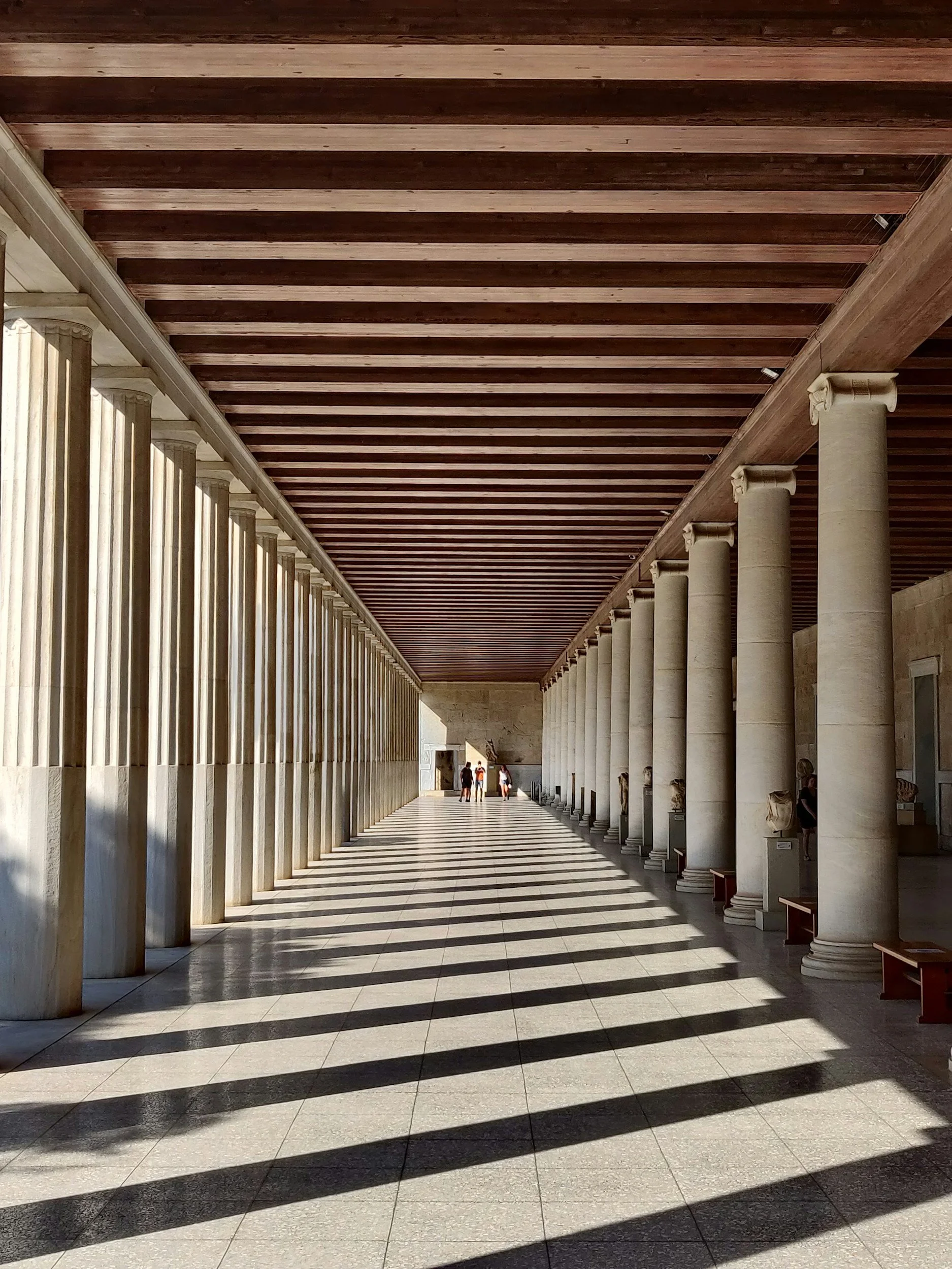Empty corridor with tall stone columns on the right, a row of columns on the left, and shadows cast on the floor from the ceiling beams, with a small group of people in the distance.