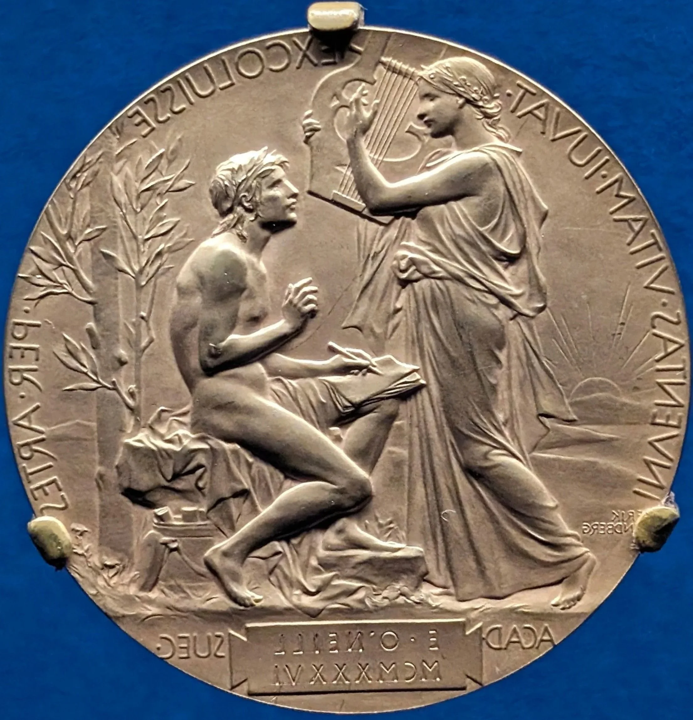 Close-up of a silver commemorative medallion featuring a seated man with a laurel wreath on his head reading a book, and a standing woman with a harp, an olive branch, and flowing drapery. The medallion has Greek inscriptions around the edge.