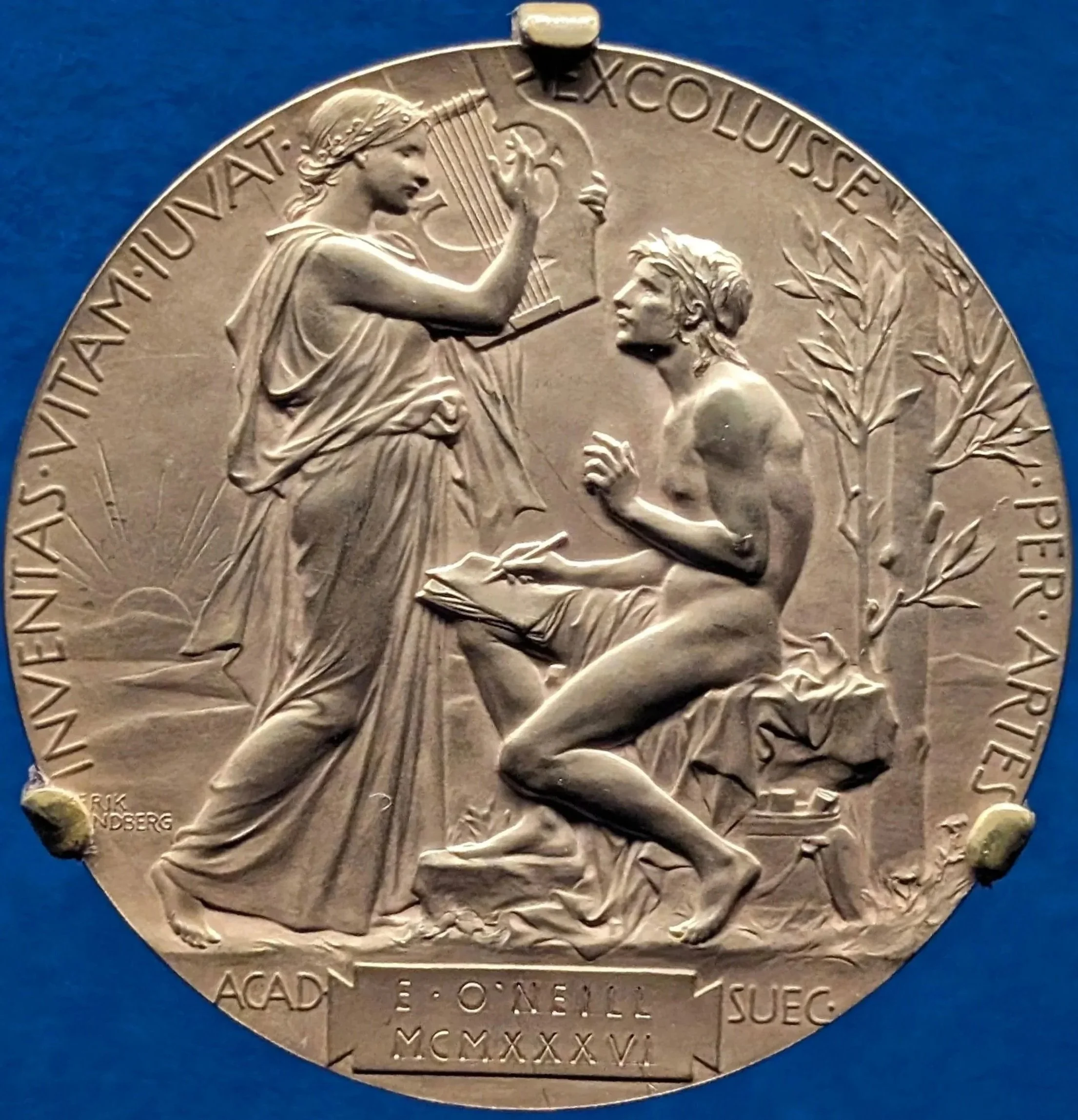A medal depicting a seated man writing in a book and a standing woman holding a torch and a tablet, with trees and a rising sun in the background. The words around the edge include Latin phrases and the year 1926.