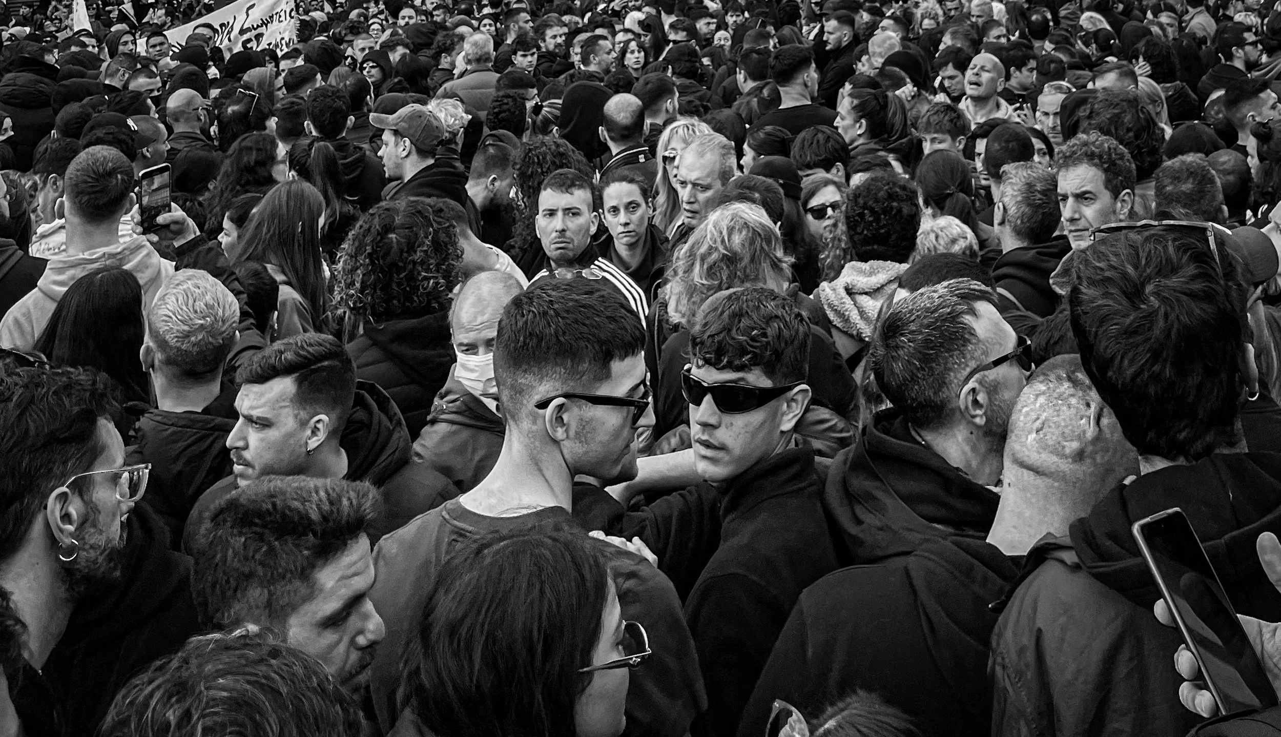 A large crowd of people gathered together at a protest or demonstration, mostly facing forward with some engaging in conversations or taking photos.