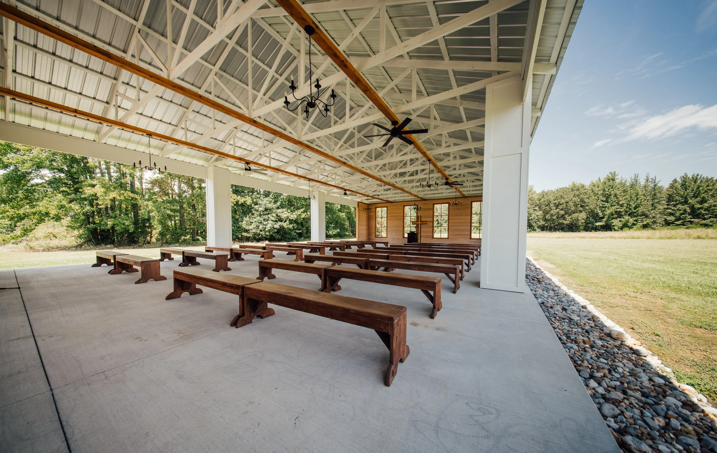 Empty outdoor church pavilion with wooden benches, a cross on the back wall, and a small altar, set in a green landscape on a sunny day.
