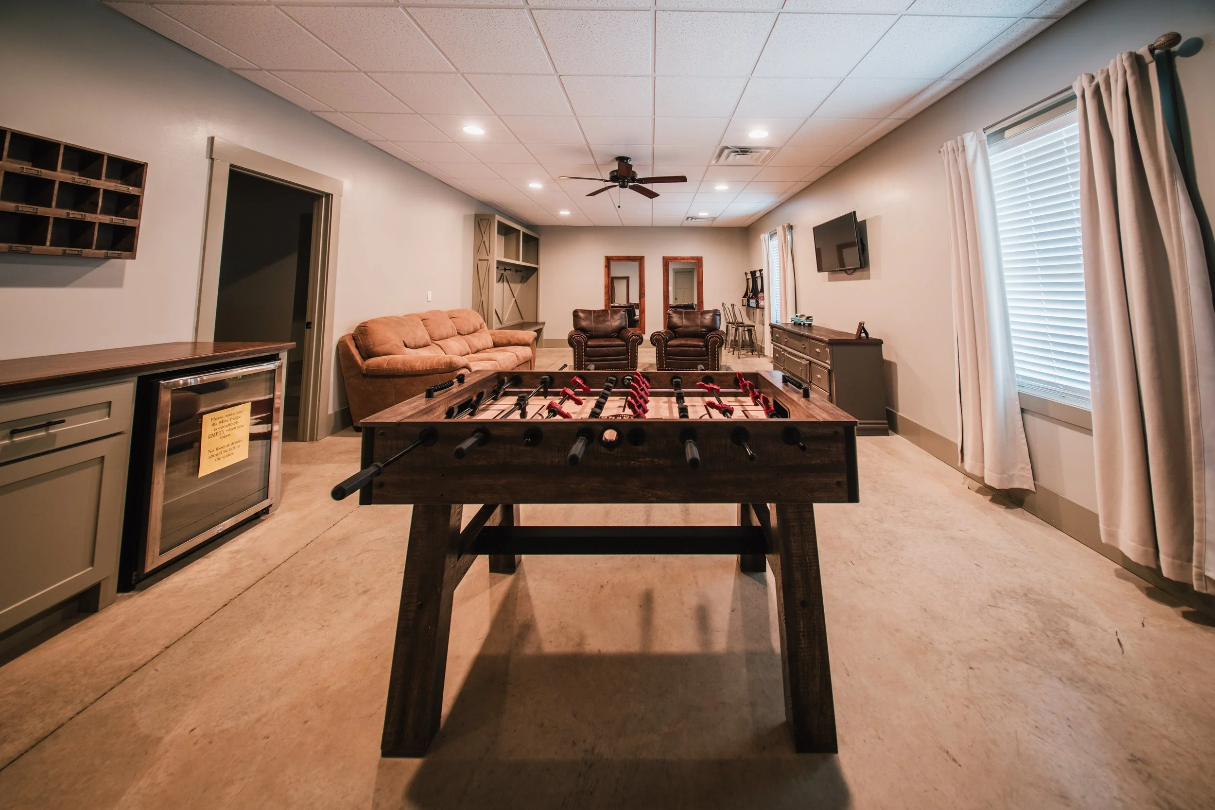 A game room with a foosball table in the foreground, a beige sofa on the left, brown armchairs facing the room, a TV on the right wall, and windows with curtains on the right side.
