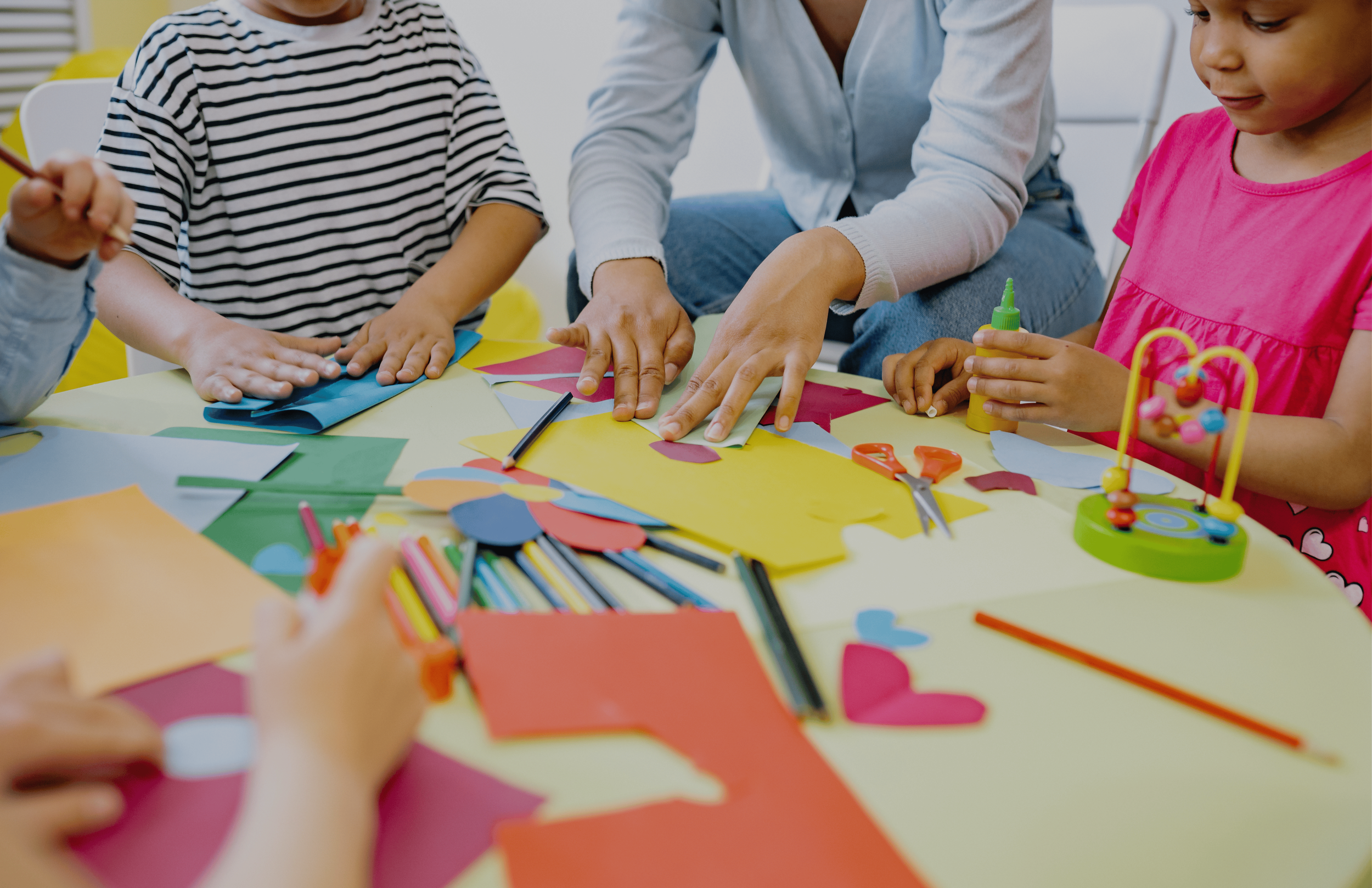 Children and an adult creating arts and crafts with colorful paper, scissors, glue, and markers at a table. Kids birthday party setup at indoor play place in Howell MI.
