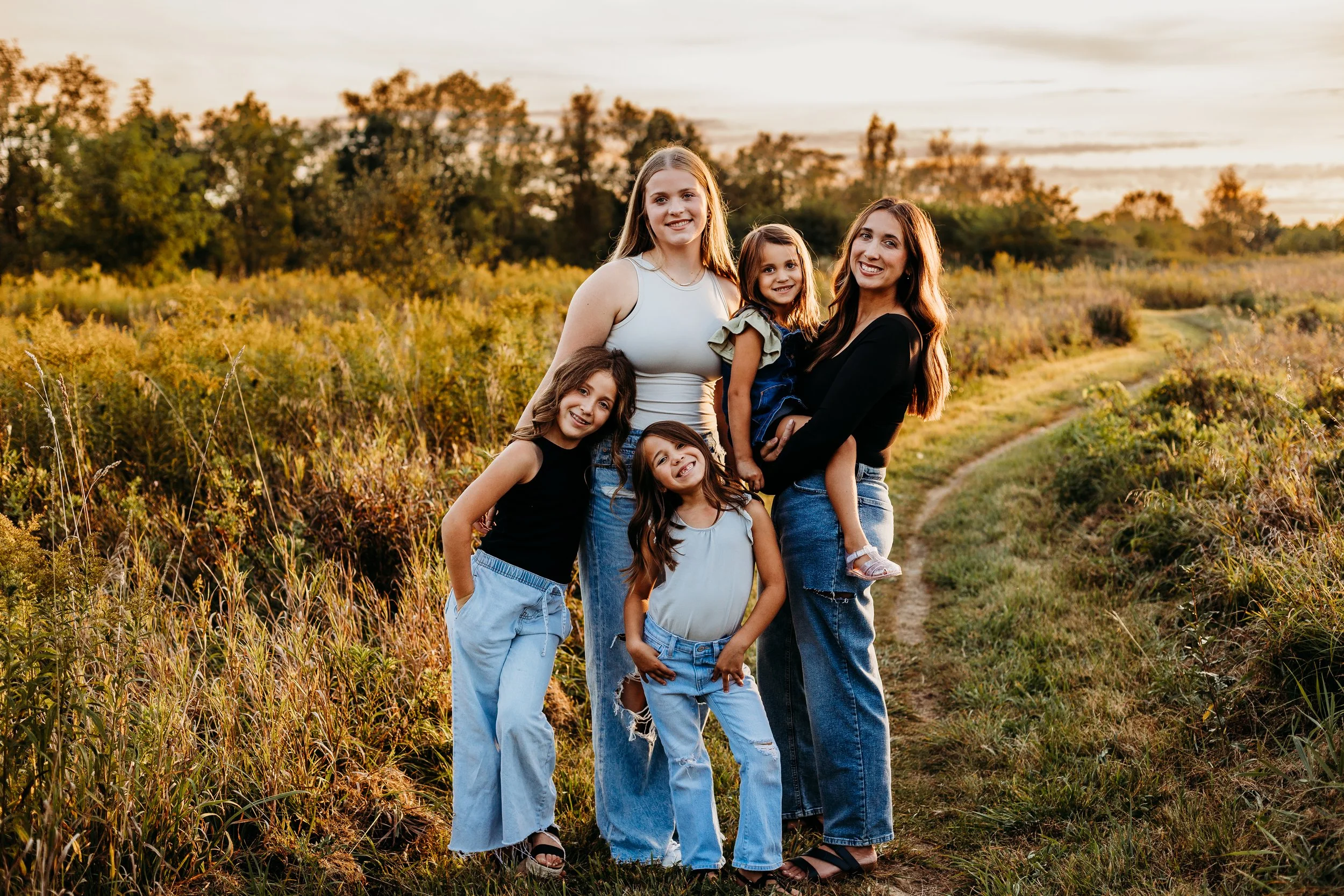 A group of five women, including three young girls and two adult women, smiling and posing together outdoors on a dirt path in a field during sunset. Safe and clean indoor play center for families in Howell MI.