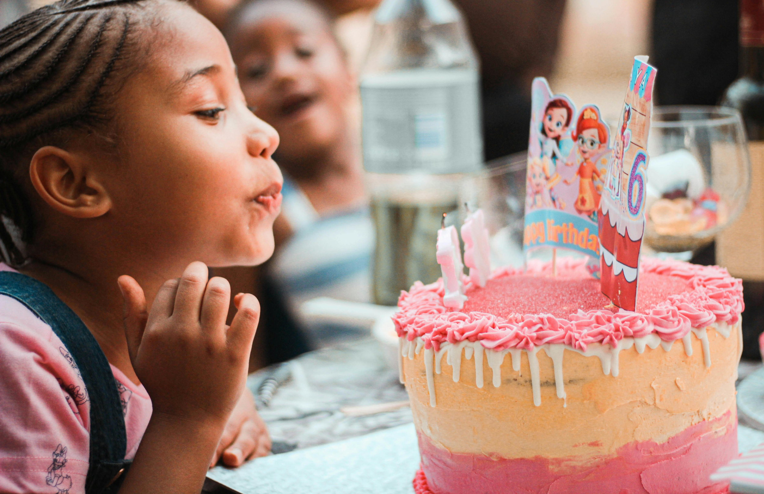 A young girl blowing out candles on a pink and white birthday cake decorated with 'Happy Birthday' and cartoon character toppers at a birthday celebration. Indoor play place in Howell MI.