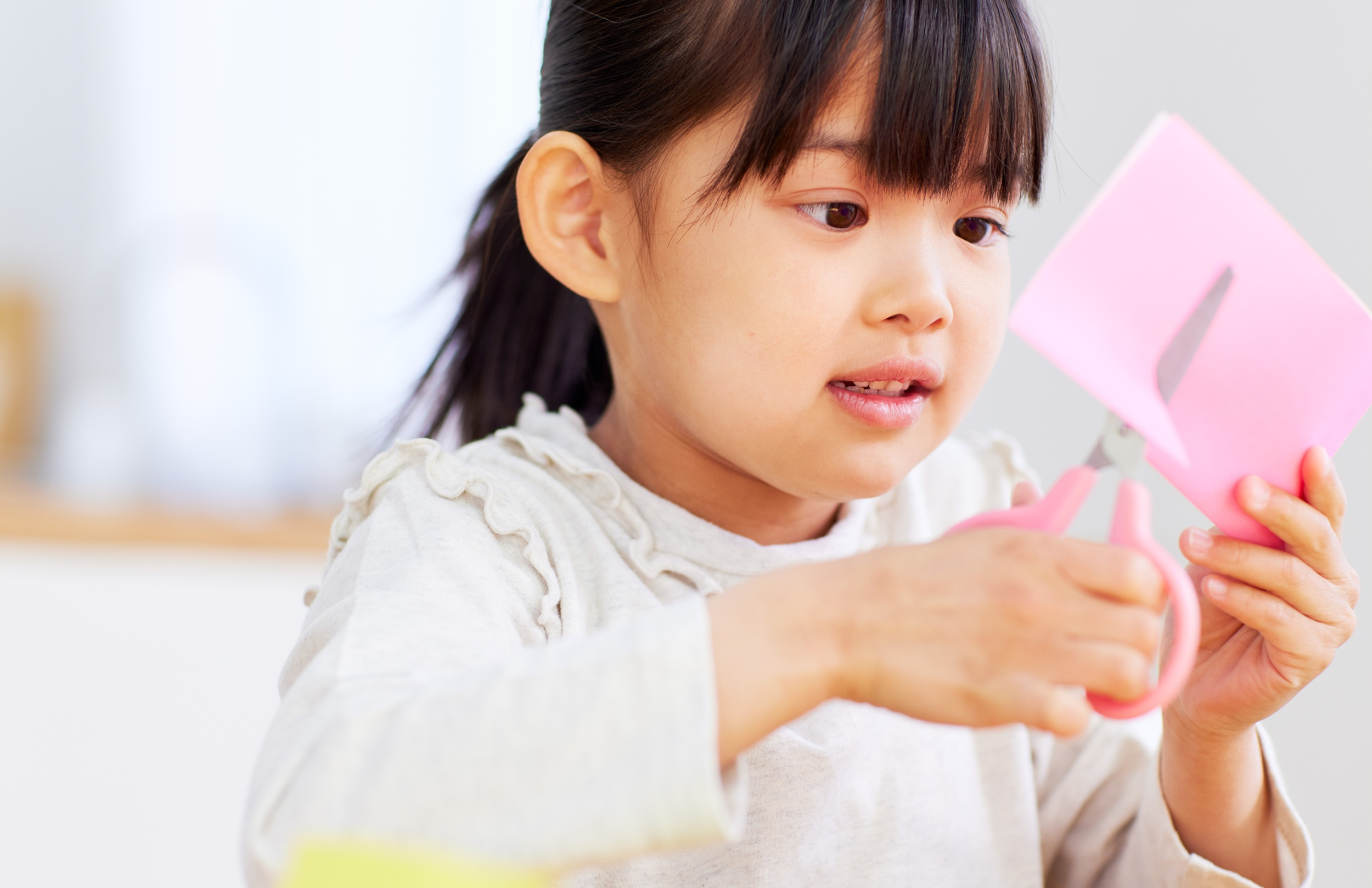 A young girl with dark hair holding pink scissors and paper, cutting a pink paper with a gray design on it. Local kids activity center near Brighton and Howell.